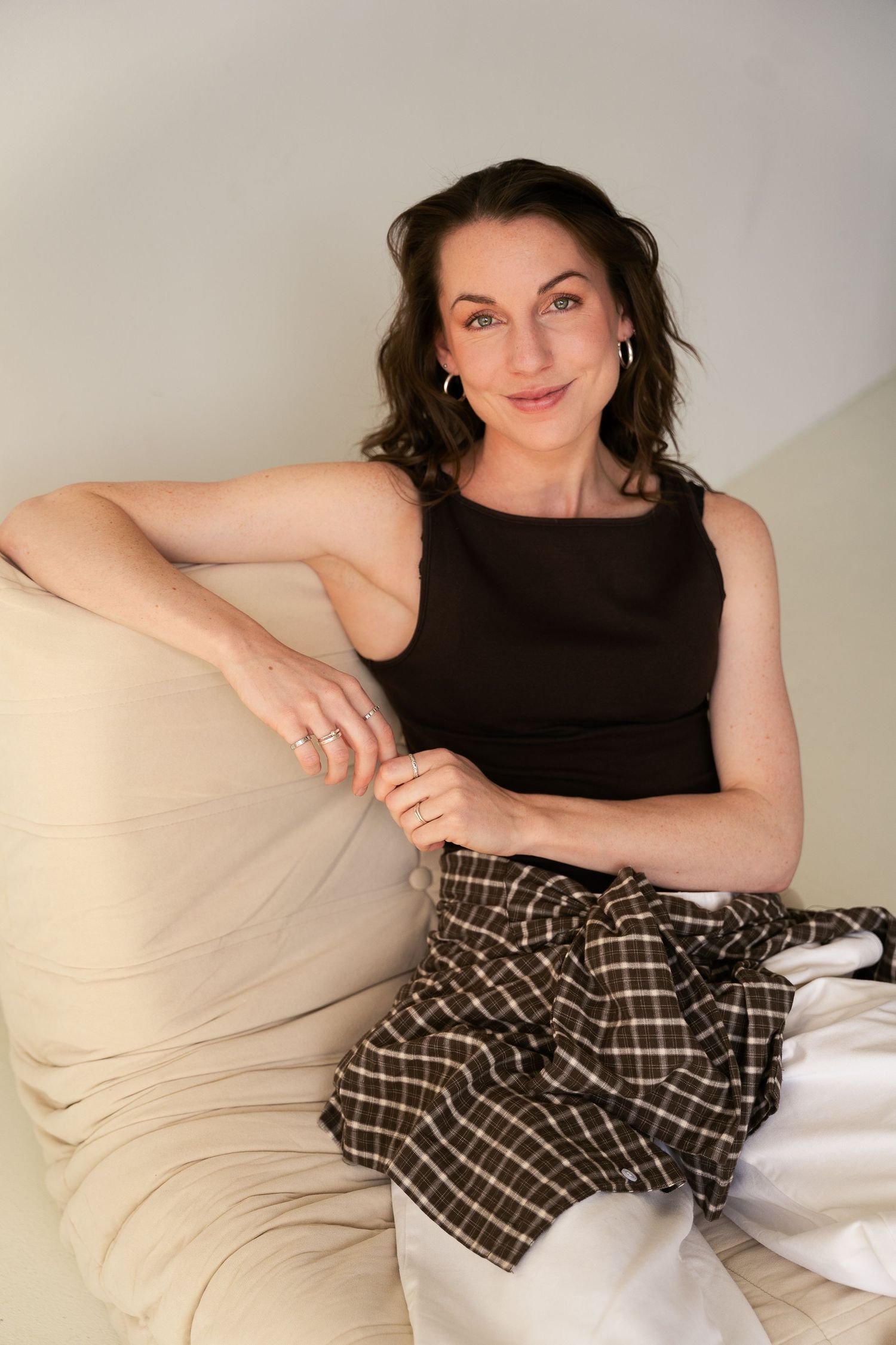 Woman in black tank top and plaid skirt sitting relaxed on beige sofa, smiling warmly at camera.