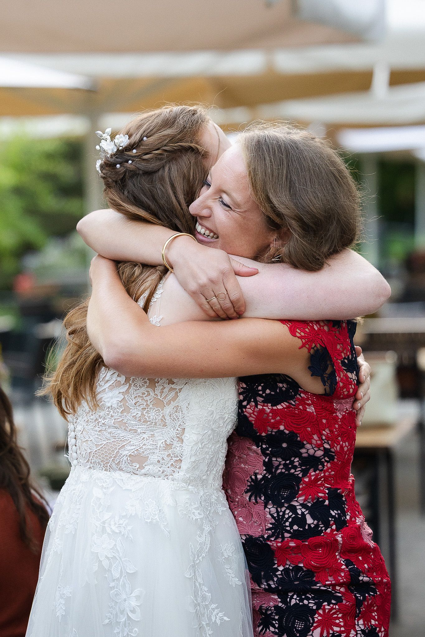 Twee mensen knuffelen elkaar op een bruiloft, de ene draagt een trouwjurk en de andere een feestelijke rode jurk. Zomerbruiloft in Rozendaal