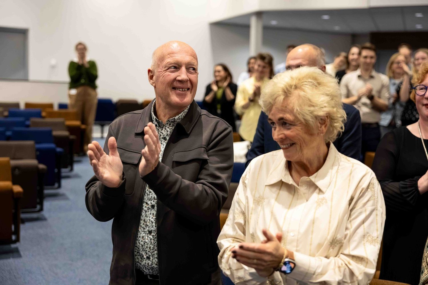 Two elderly people clapping and smiling at an indoor event, surrounded by a seated audience in a modern auditorium.