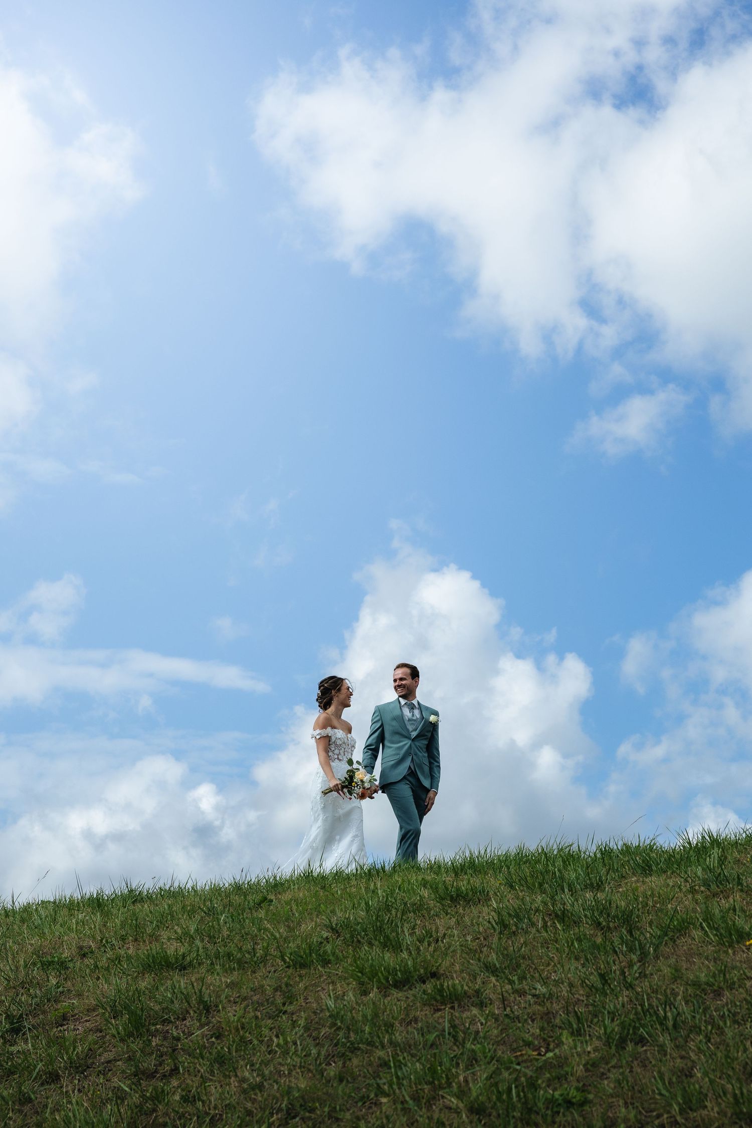 Bride and groom holding hands on a grassy hill under a bright blue sky with white clouds on their wedding day.