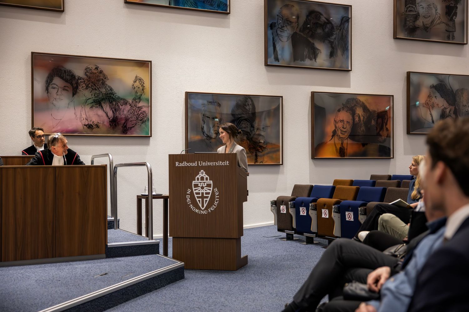 Speaker at Radboud University podium addressing audience in lecture hall with colorful paintings on wall.