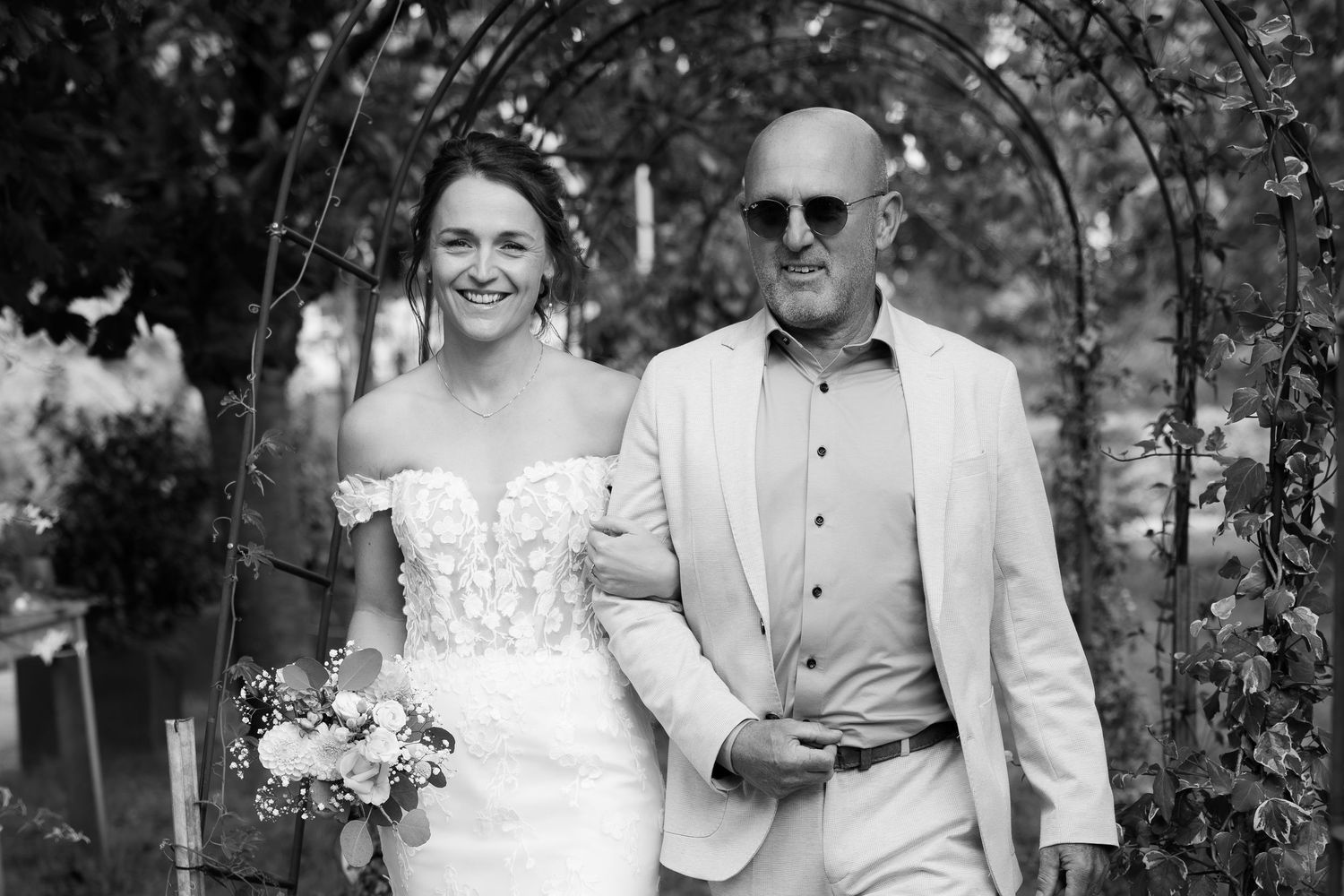 Black and white photo of a smiling bride in a strapless lace gown walking with a man in a suit under a floral arch.