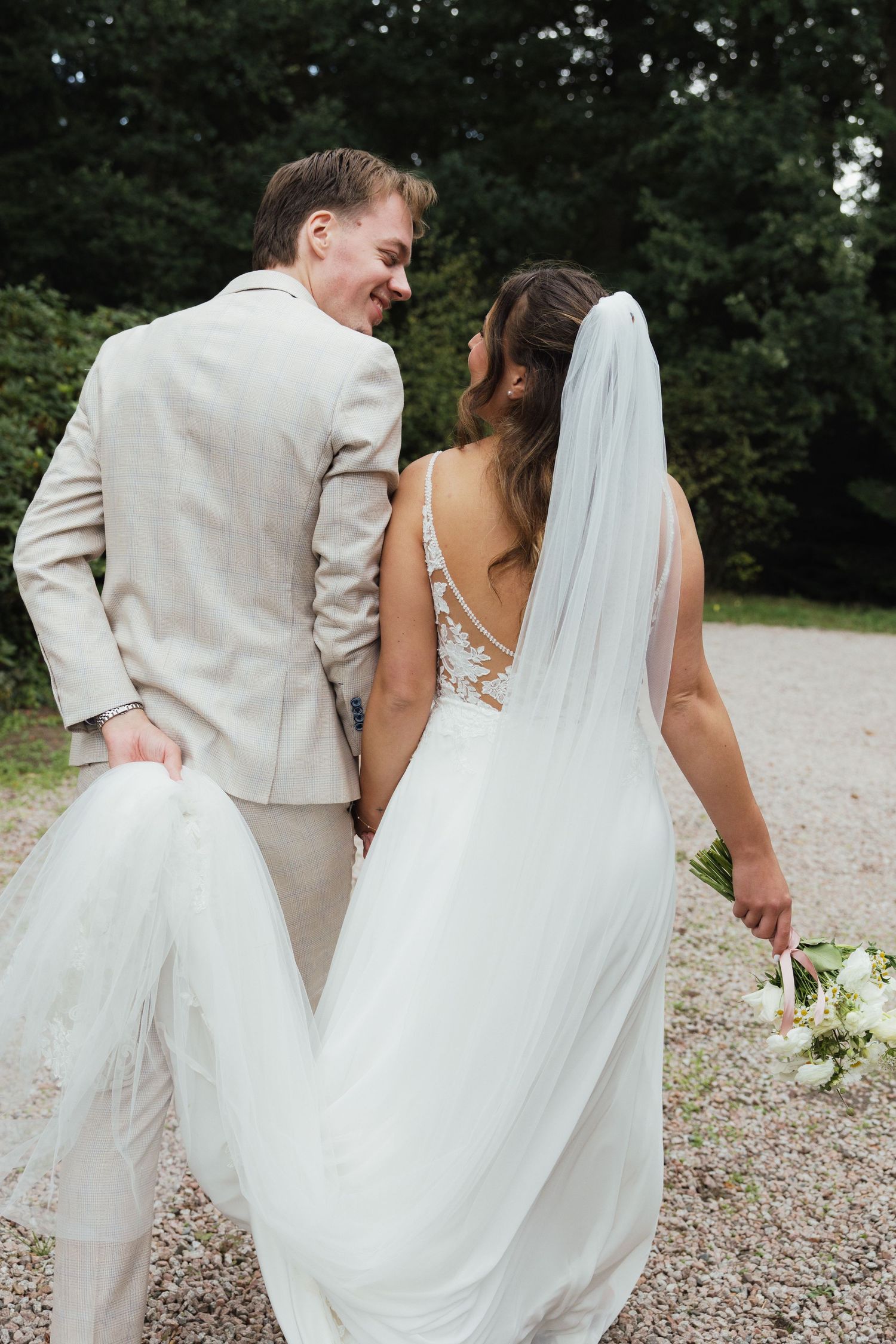 A smiling groom in a beige suit walks with his bride in a white lace gown and veil, holding a bouquet outdoors.