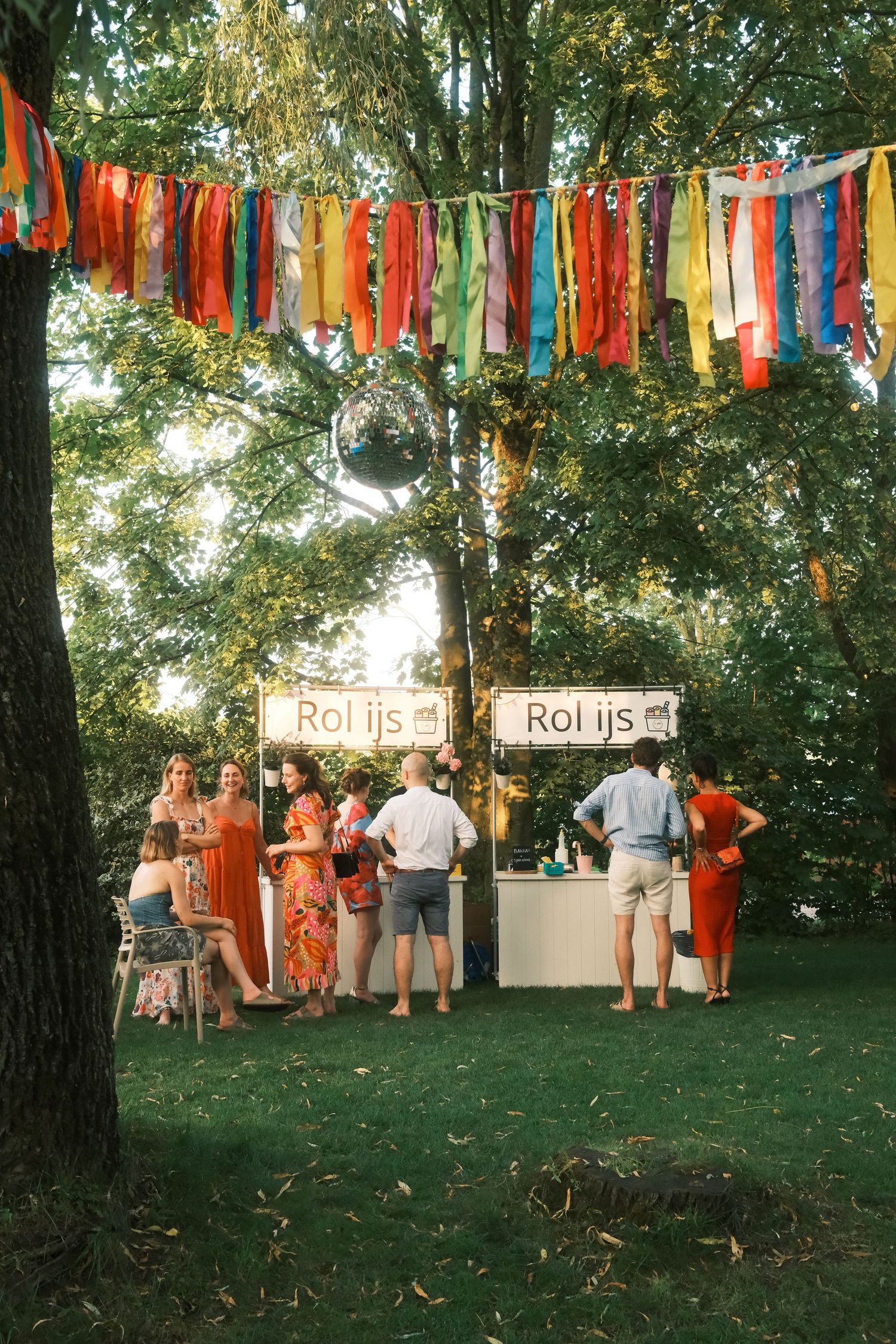 Outdoor garden party with colorful ribbon bunting, disco ball, and guests enjoying rolled ice cream stands among lush trees.