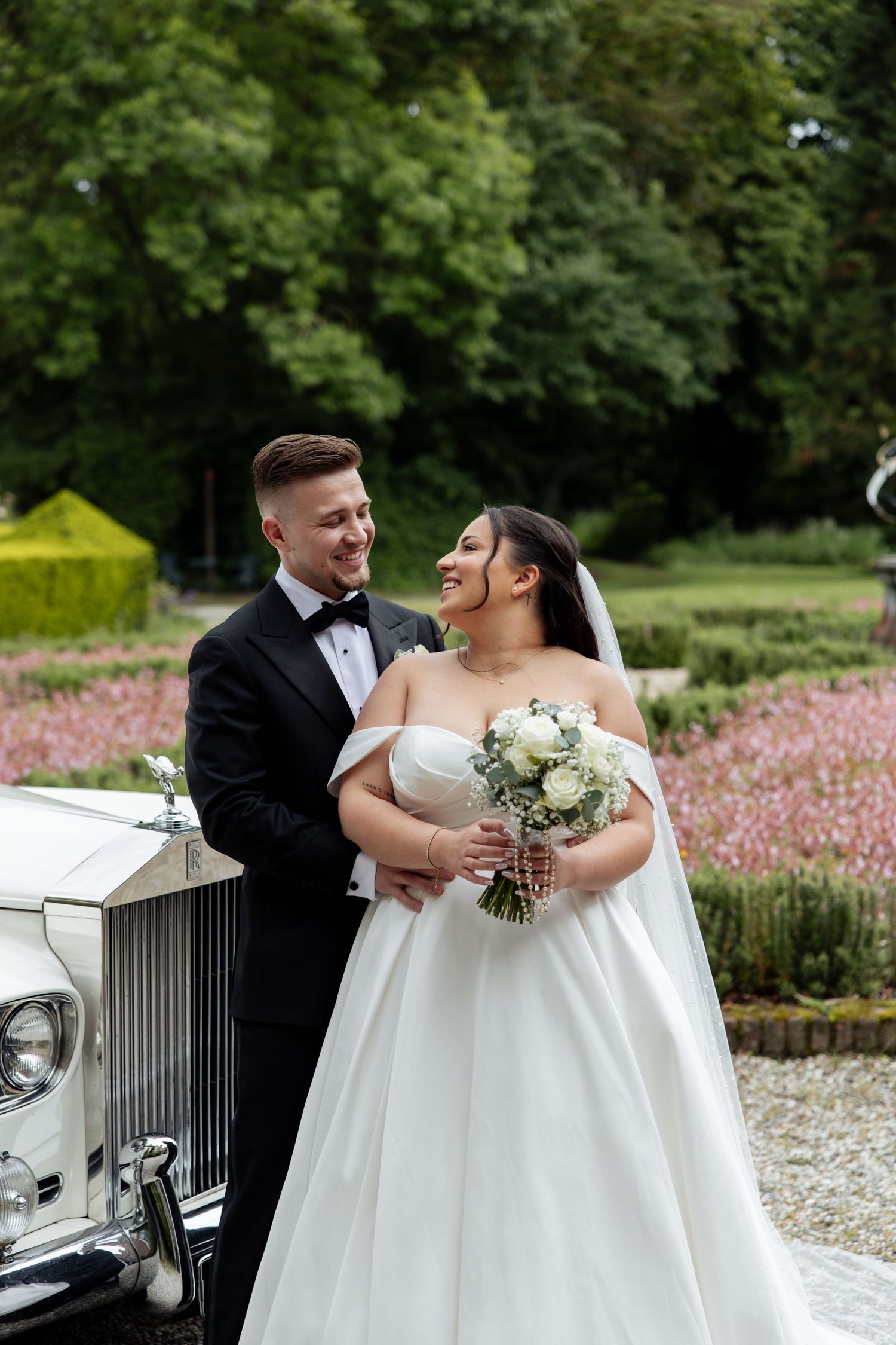 Bride and groom smiling together outdoors by a vintage white car, surrounded by lush gardens on their wedding day.