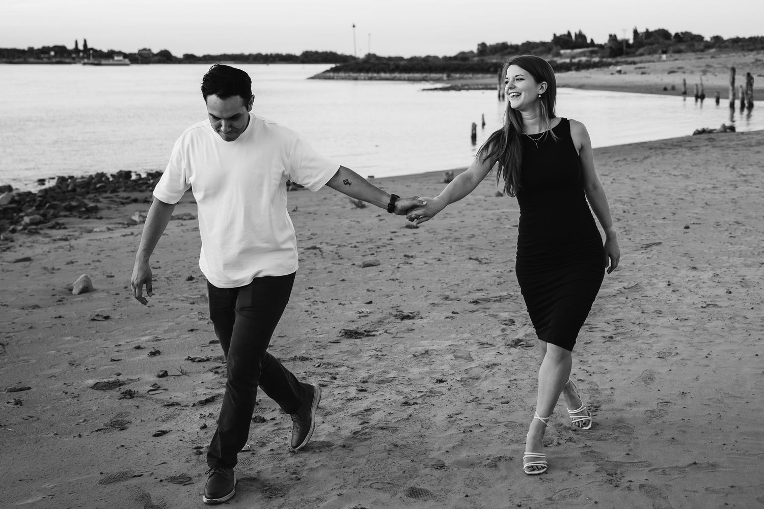 Black and white photo of a couple holding hands and walking on a sandy beach at sunset.