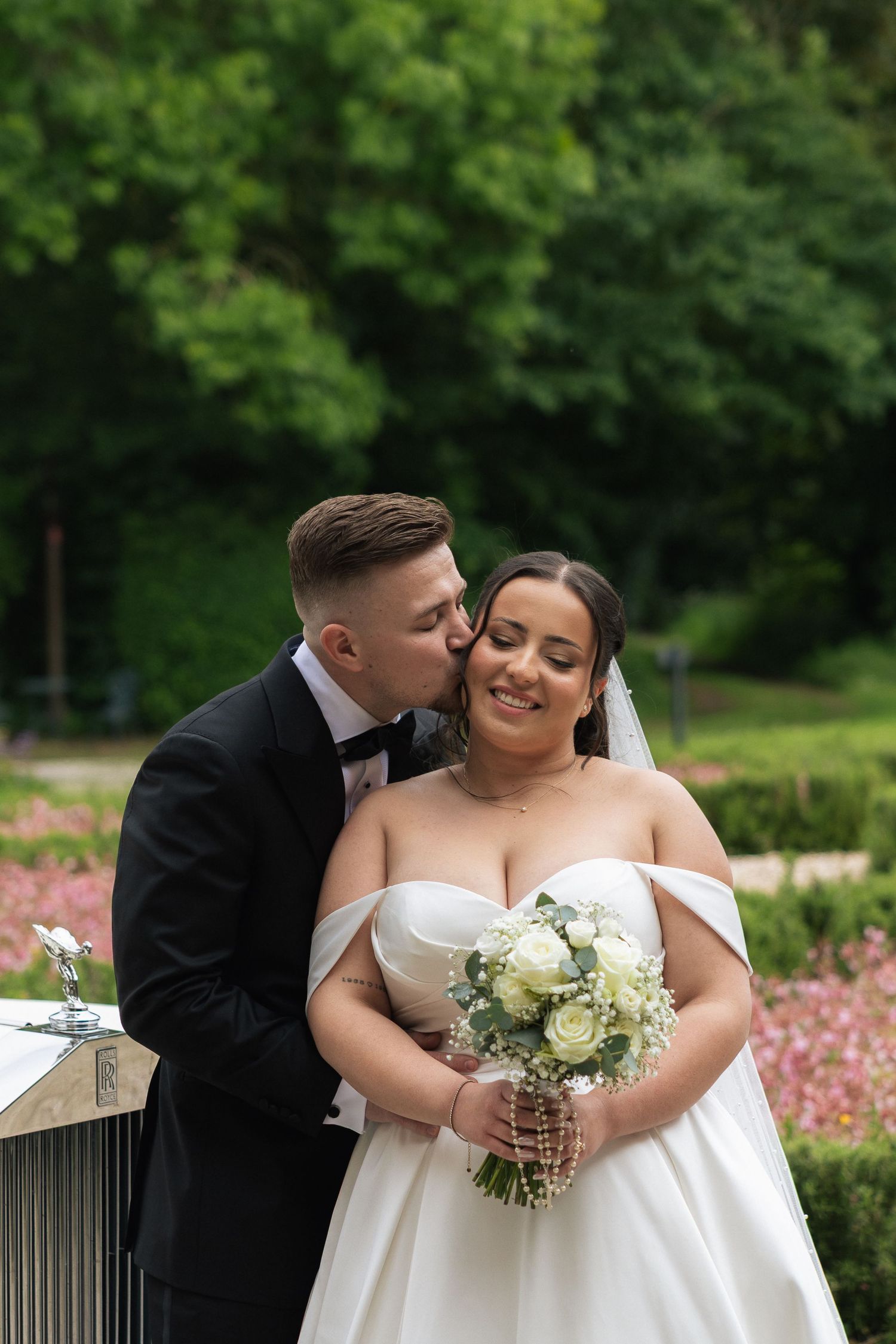 Groom in black tuxedo kissing bride on cheek as she smiles holding white rose bouquet in lush garden setting.