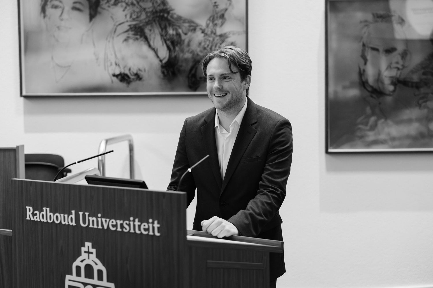 Man speaking at Radboud Universiteit podium in black and white photo, smiling in formal attire.