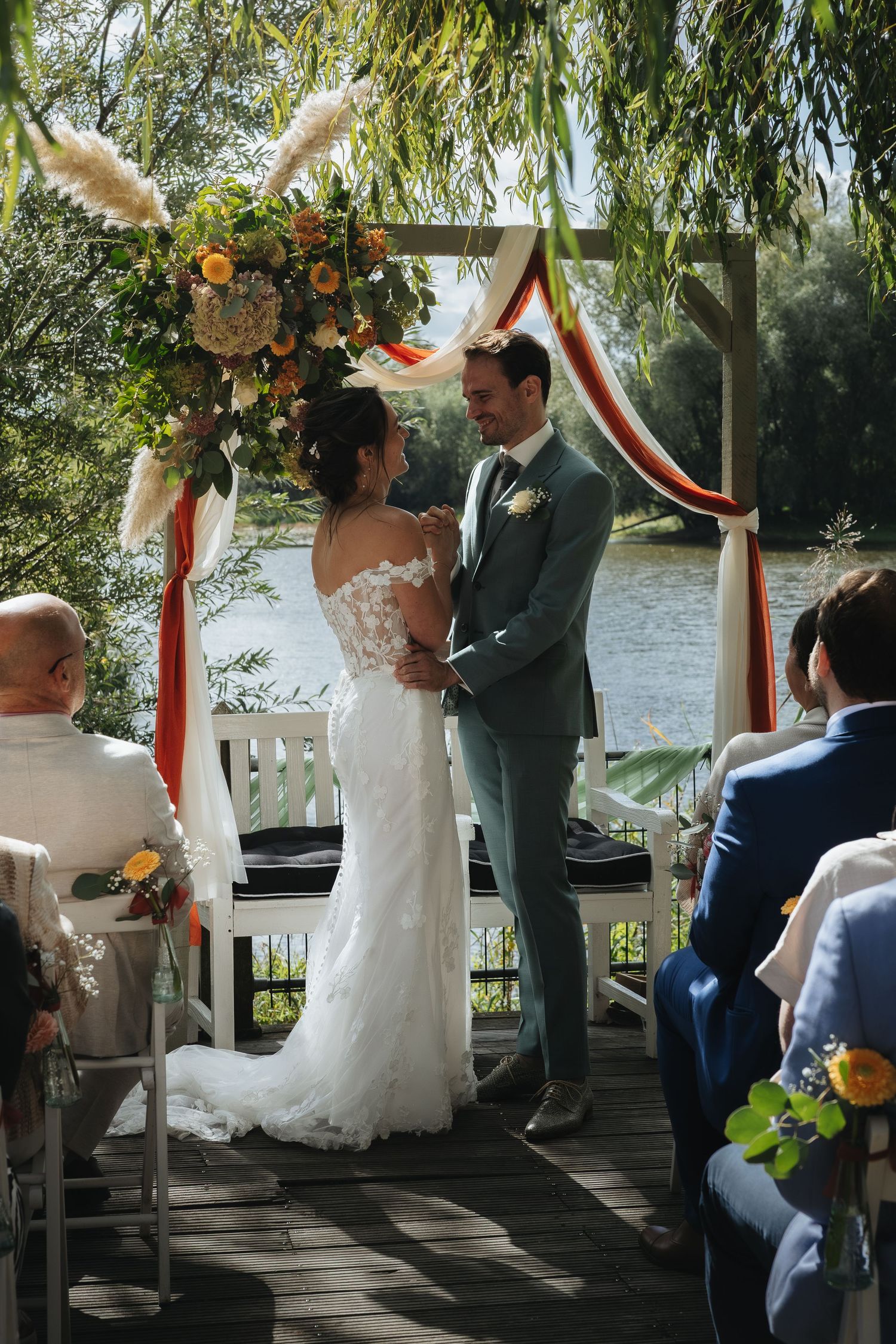 Bride and groom exchange vows under floral arch by lake at outdoor wedding ceremony.