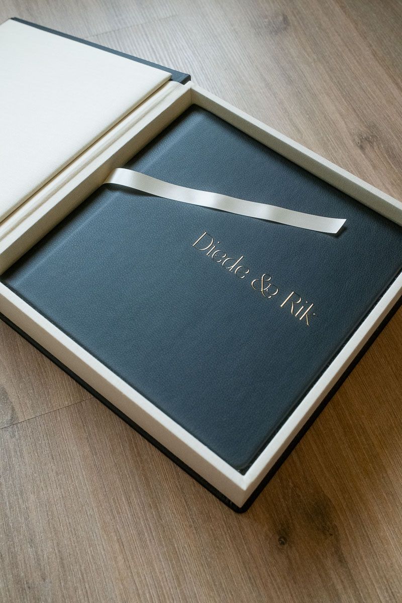 Elegant dark gray wedding photo album with gold names and ribbon, presented in a luxury keepsake box on wood floor.