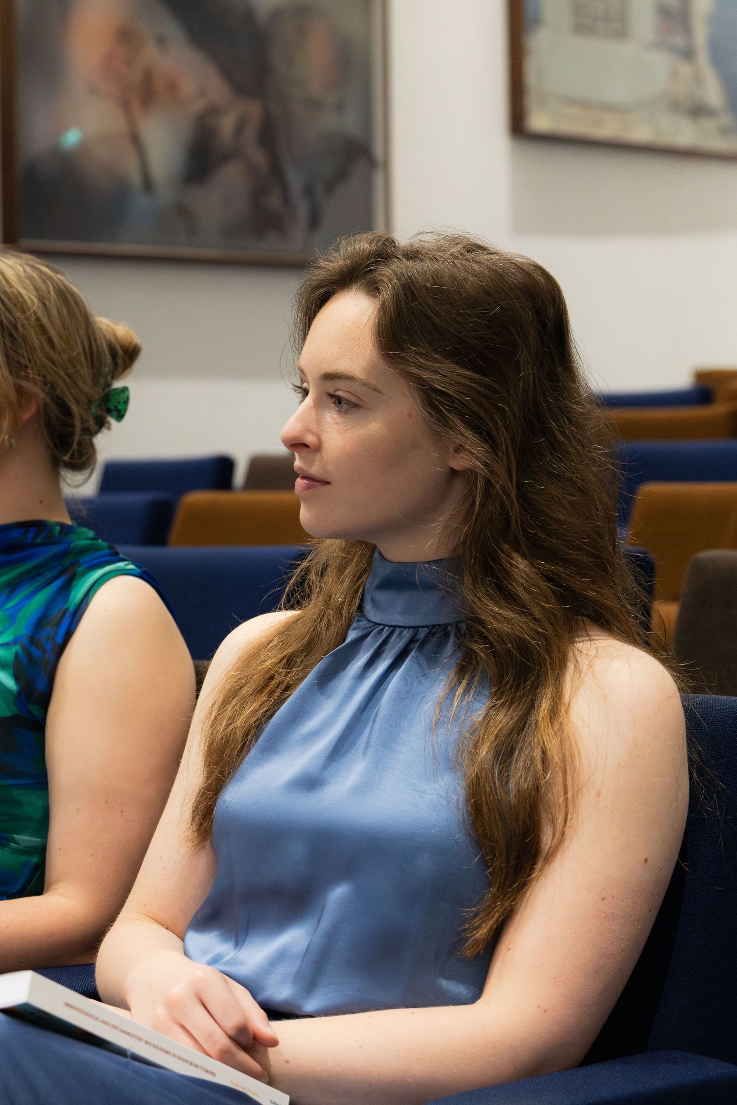 Young woman in blue sleeveless top sitting in conference room chairs, holding a book, looking attentively to the side.