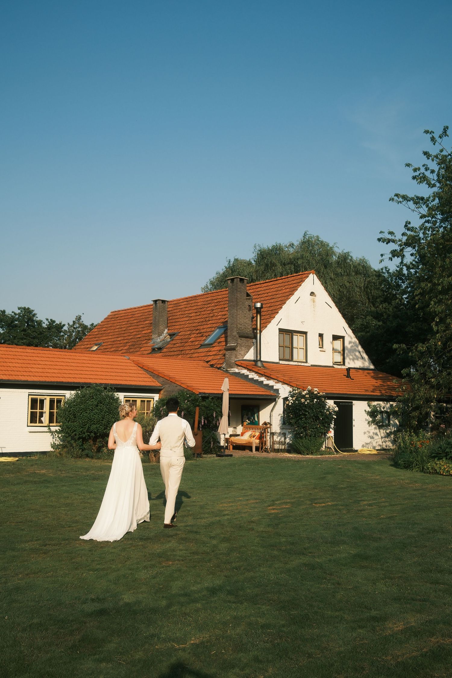 Bride and groom walking hand in hand across green lawn toward white farmhouse with red tile roof on sunny day.