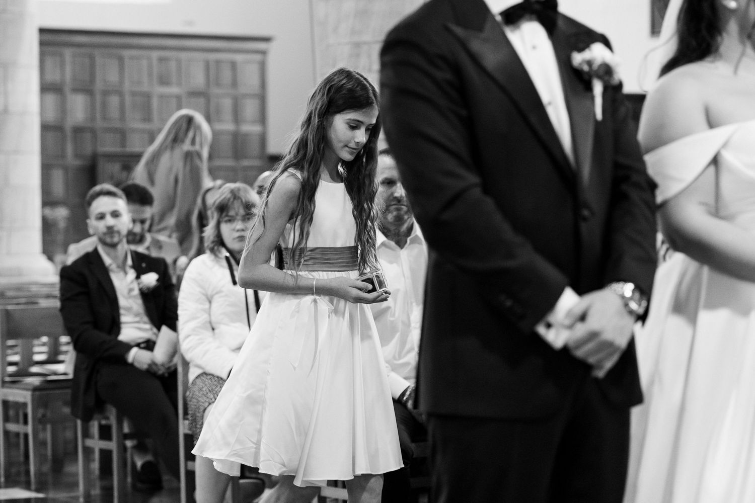 Black and white wedding photo of flower girl in white dress carrying a wooden box down the aisle.