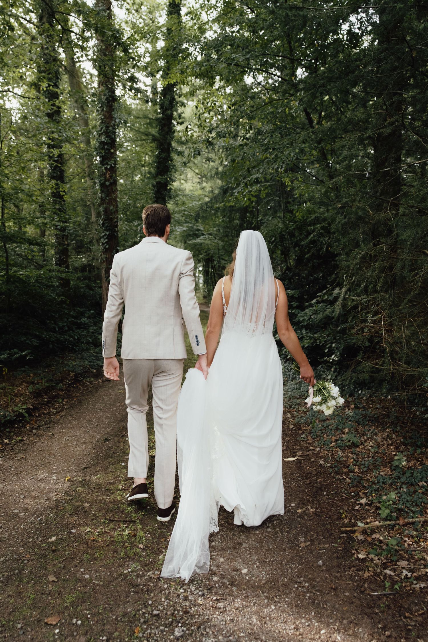 Bride and groom walking hand in hand down a forest path, bride in white gown with veil holding bouquet.