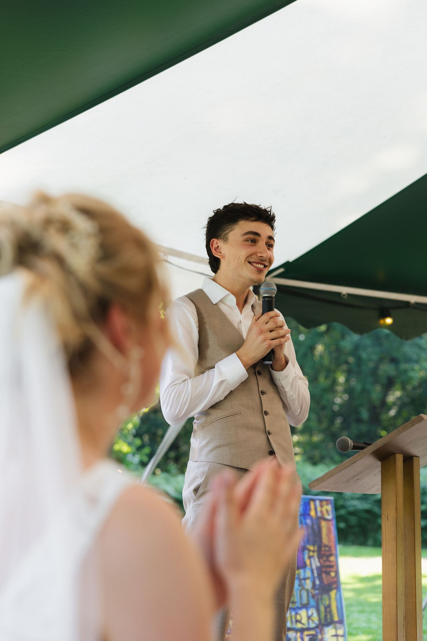Groom in grey vest speaks at outdoor wedding ceremony while bride in veil listens and claps.
