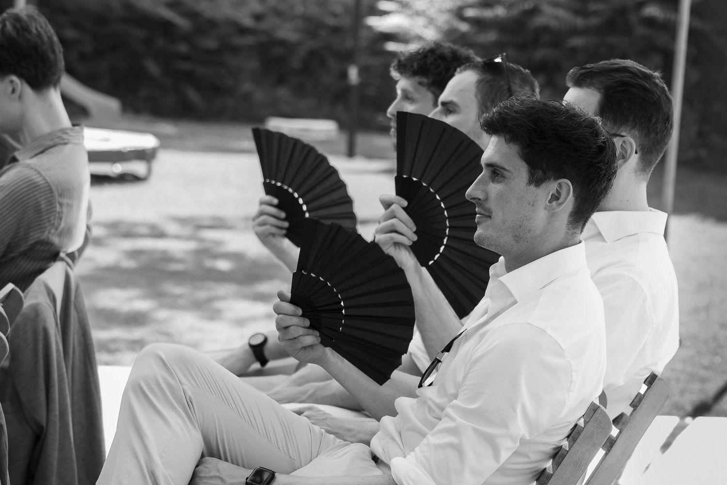 Black and white photo of men sitting on a bench holding black folding fans outdoors at a formal event.