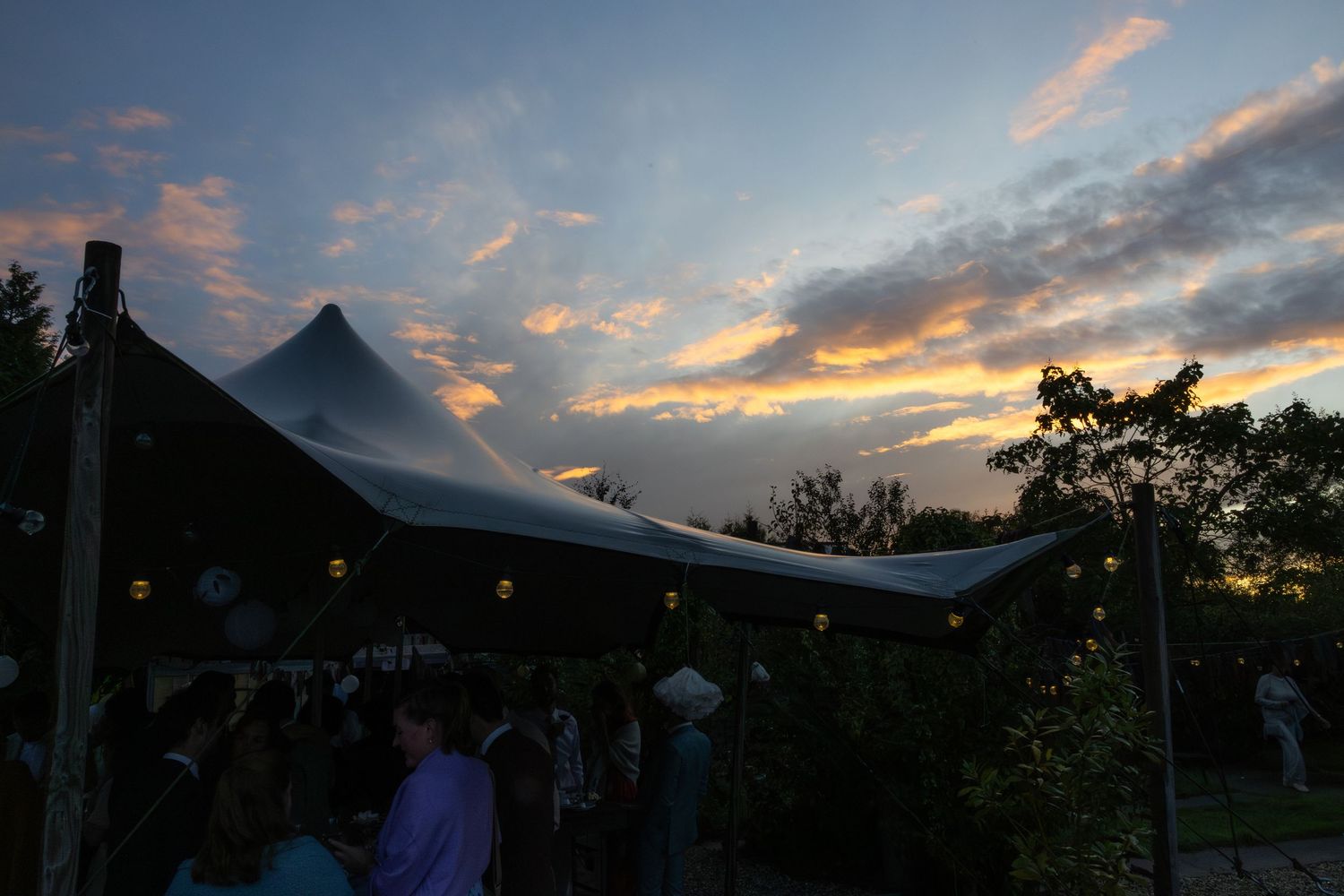 Outdoor evening event under a white tent with string lights at sunset with colorful clouds in the sky.