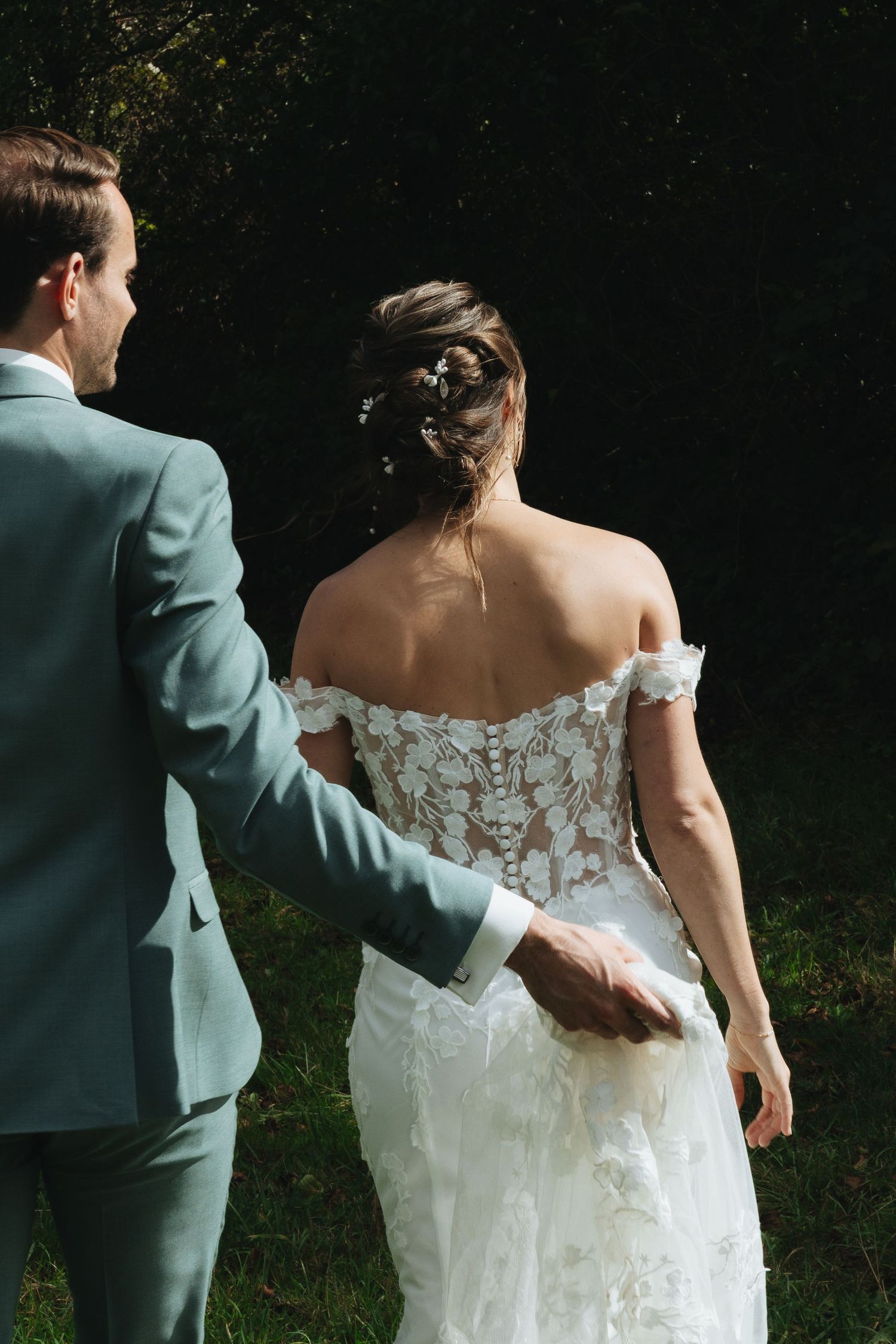 Groom in grey suit escorts bride in lace off-shoulder wedding dress walking toward dark green trees.