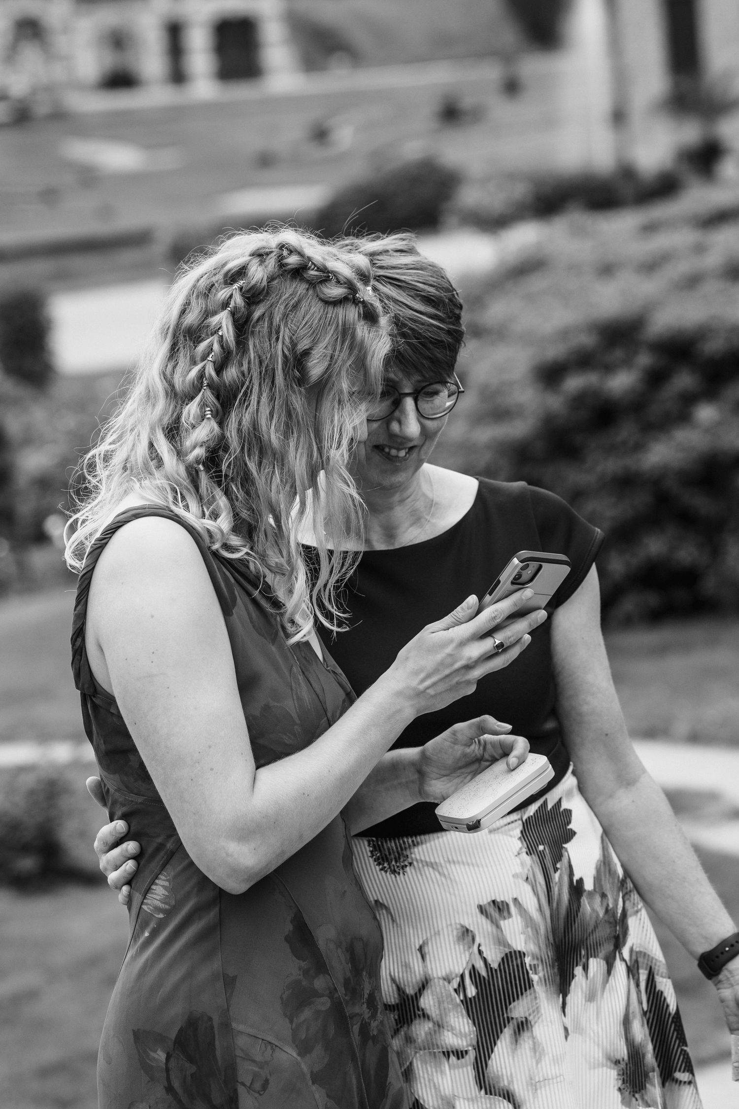 Two women smiling and looking at a smartphone outdoors in a black and white photo.