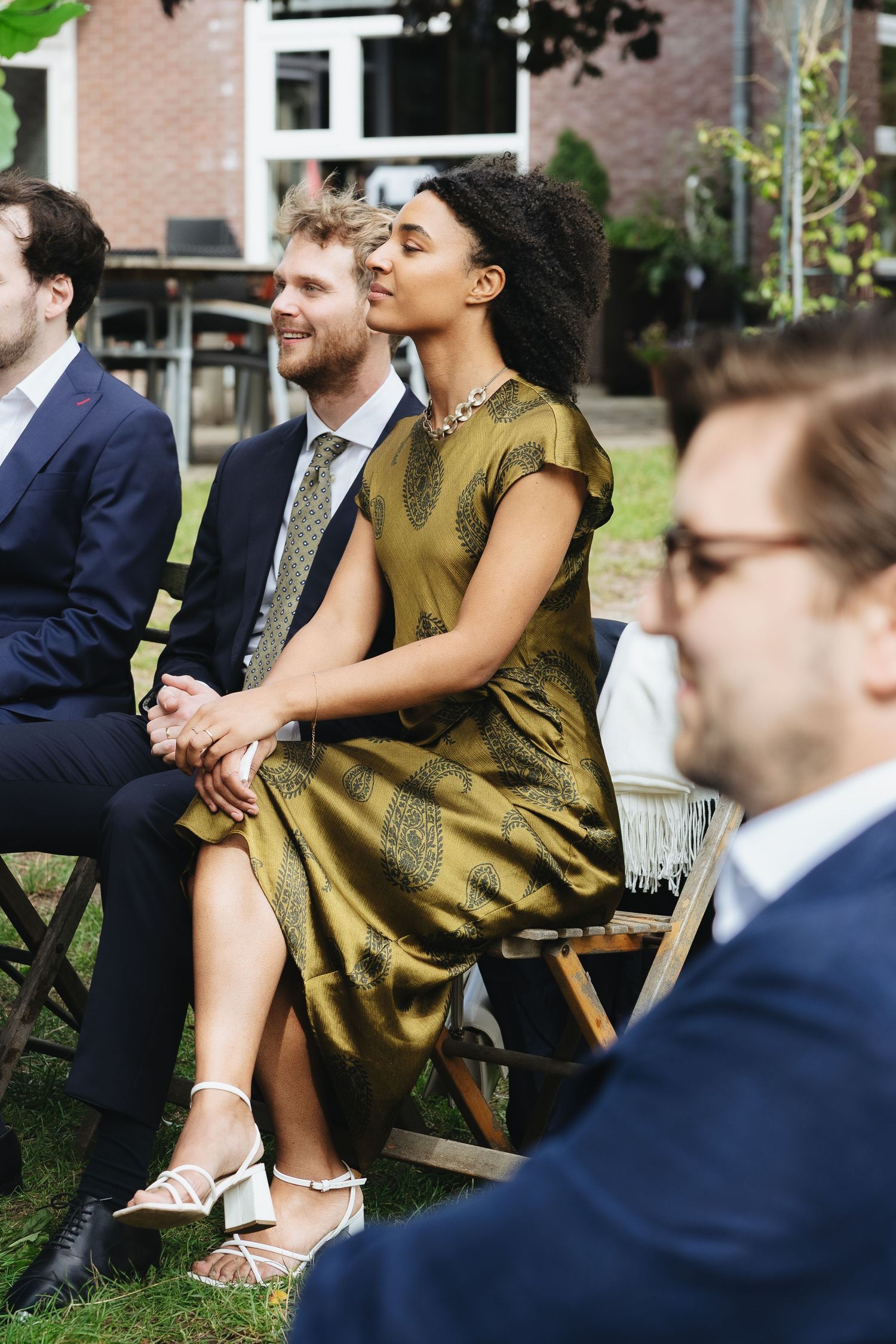 Diverse wedding guests seated outdoors, woman in gold dress and man in suit smiling during garden ceremony.