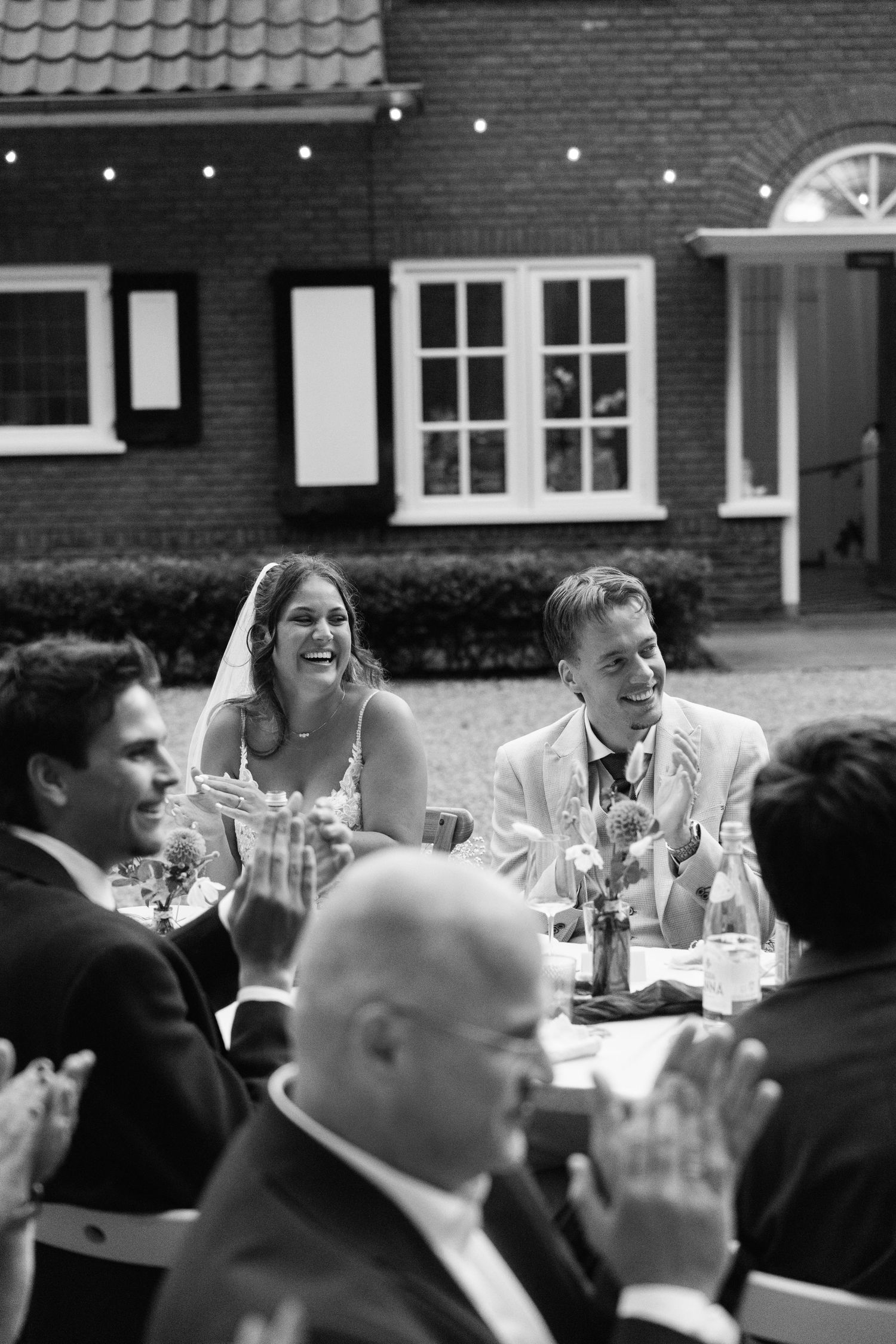 Black and white photo of a laughing bride and groom seated at outdoor wedding reception table with guests applauding.
