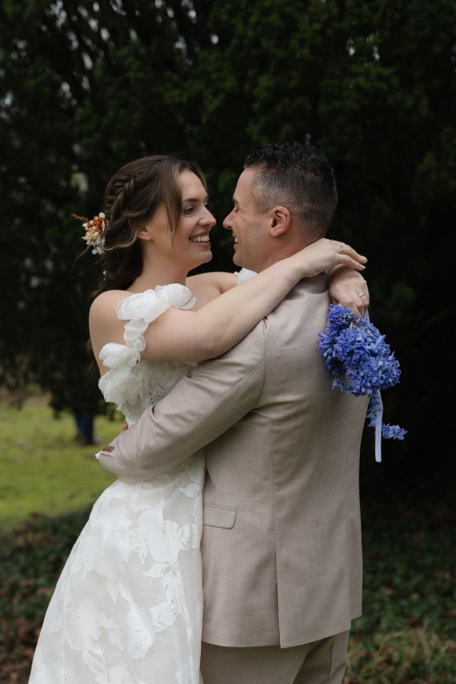 Een romantische foto van een bruidspaar dat een ongedwongen, intiem moment deelt en elkaar knuffelt. De bruid houdt een blauw boeket vast