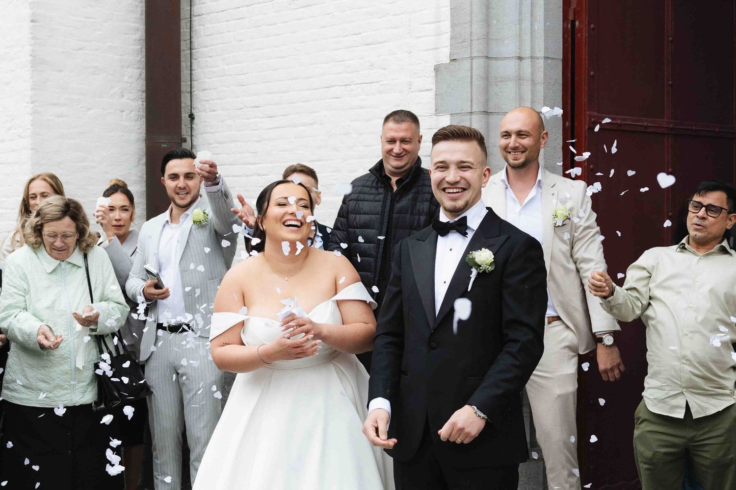Happy newlyweds exit church ceremony as guests toss white confetti in joyful outdoor wedding celebration.