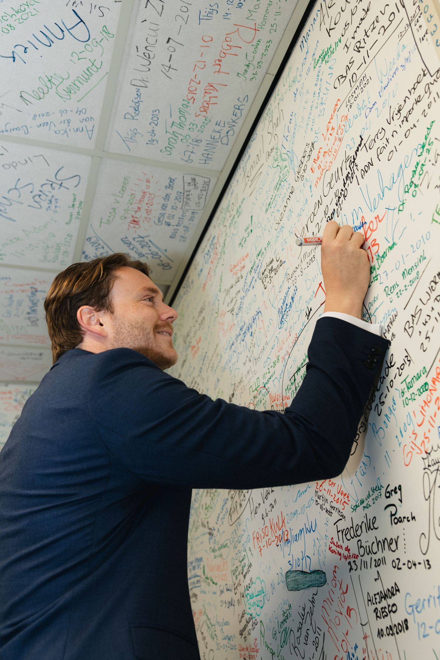 Man in blue suit signing a large white board covered in colorful signatures and messages.