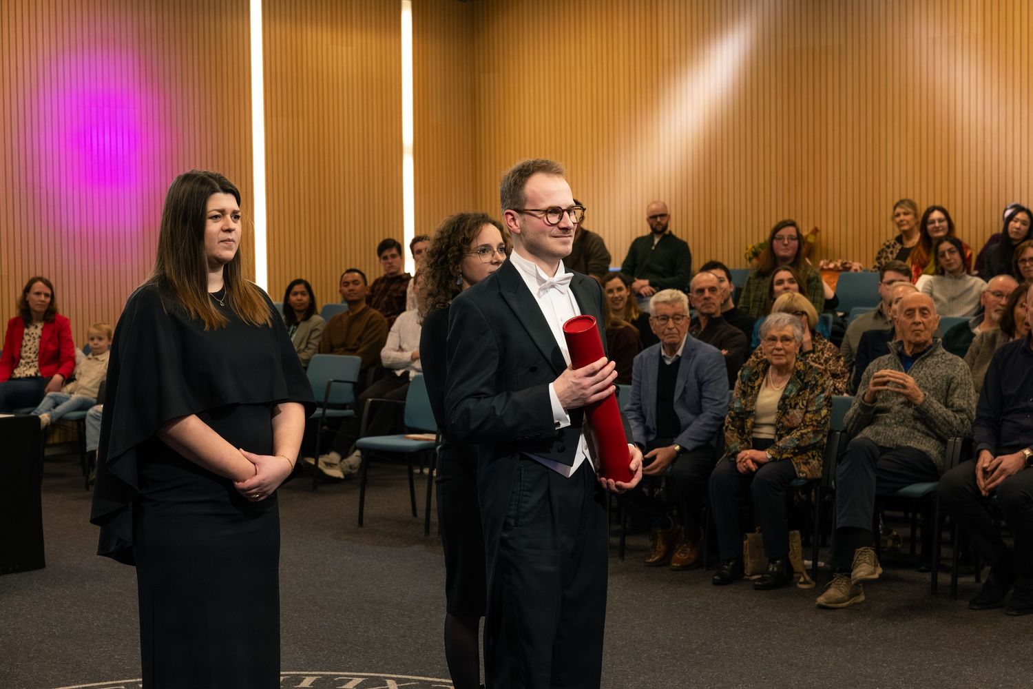 Two presenters stand before a seated audience in a modern hall, one holding a red award cylinder.