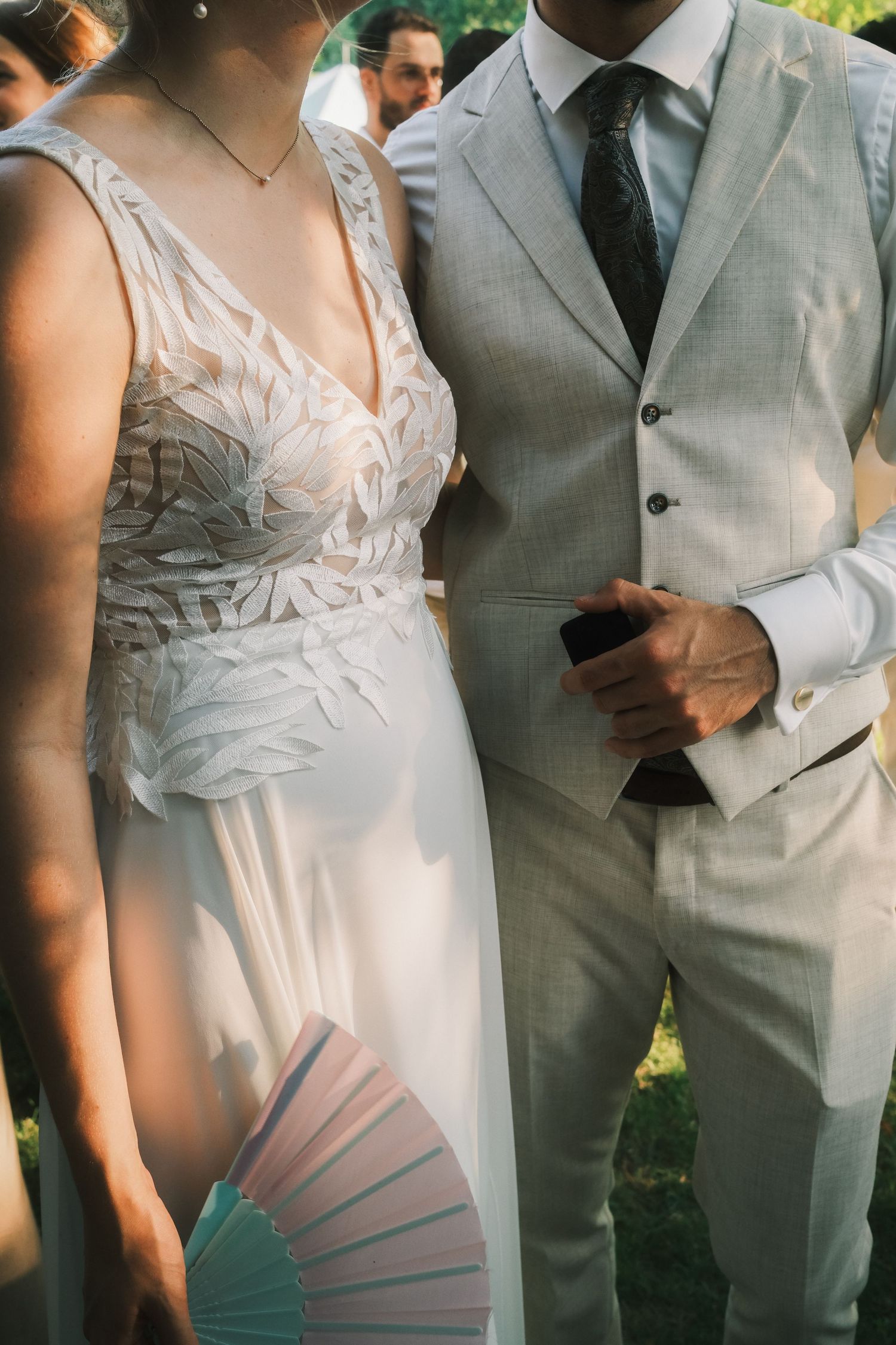 Bride in white feathered gown holding pastel fan stands beside groom in light gray suit at outdoor wedding ceremony.