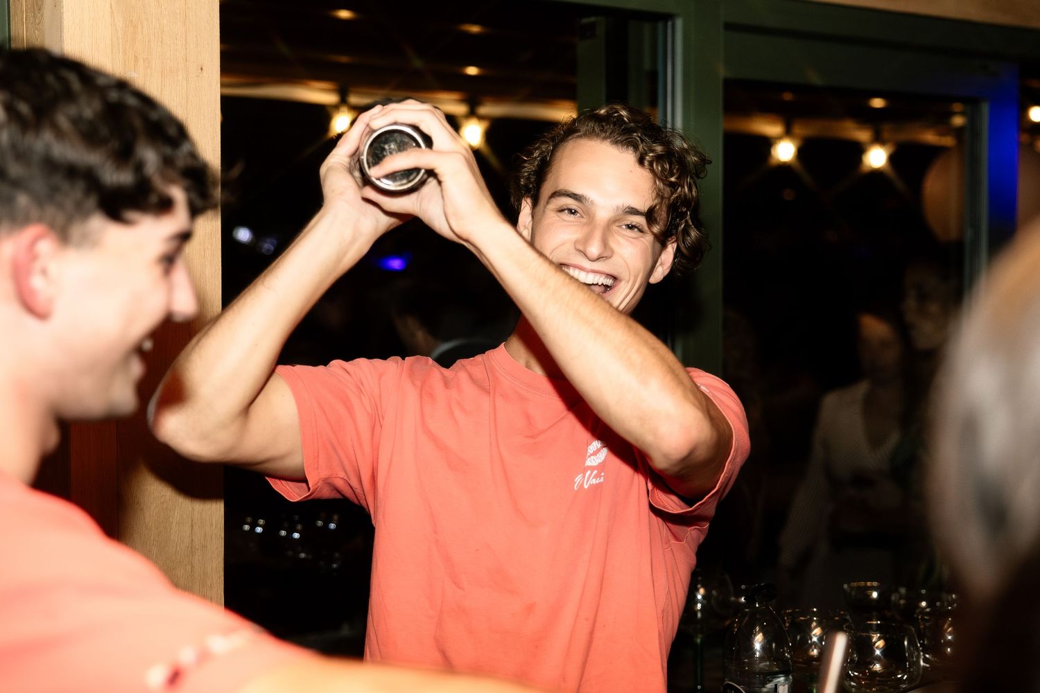 Smiling young bartender in coral shirt shaking a cocktail mixer at a lively bar event.