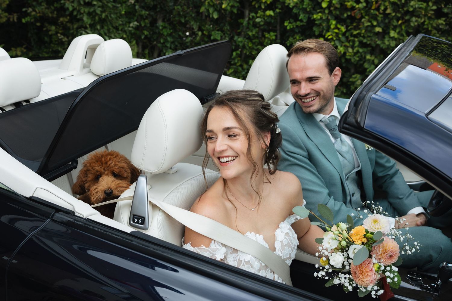 Bride and groom smiling in a convertible car with a brown dog, bride holding a floral bouquet on their wedding day.