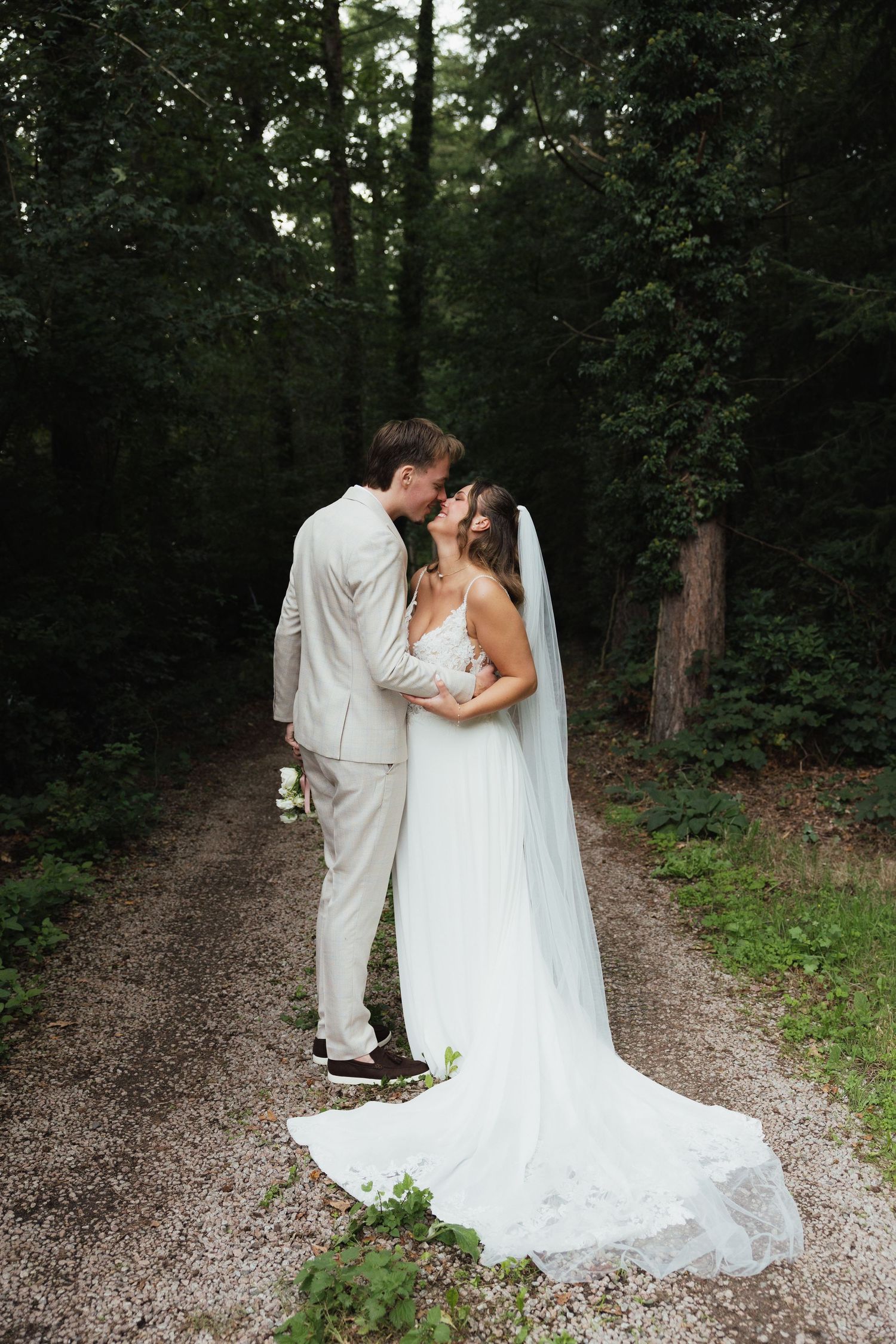 A bride and groom share a romantic kiss on a gravel path surrounded by lush green trees in a forest setting.