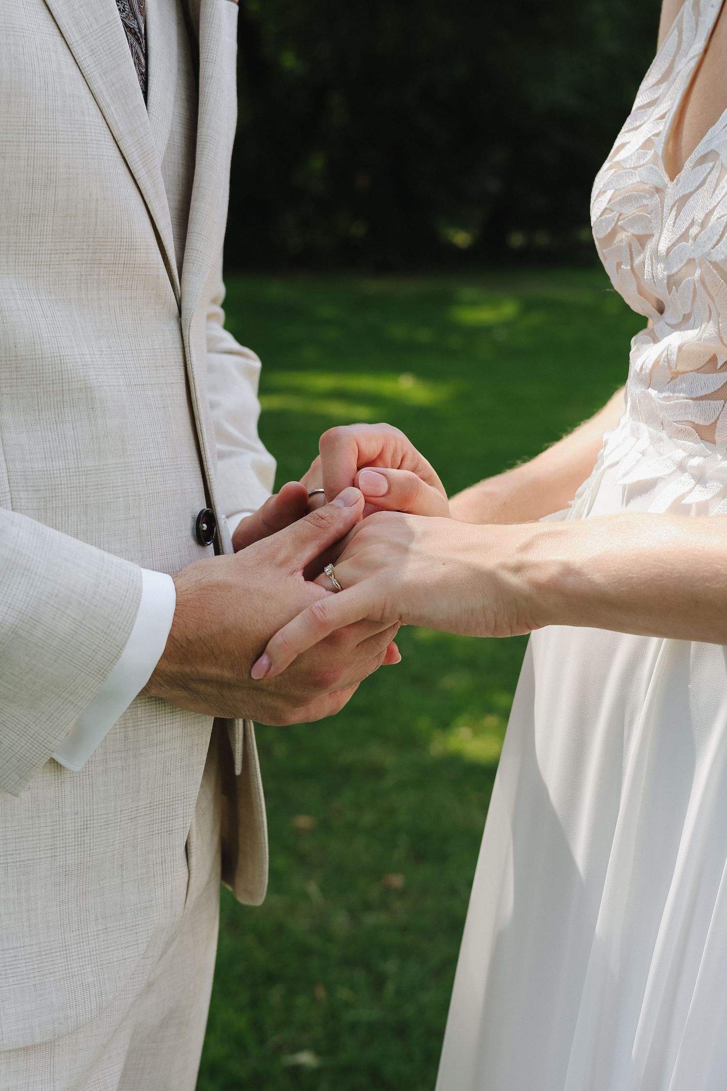 Bride placing a wedding ring on groom's finger during outdoor ceremony, both dressed in white and beige attire.
