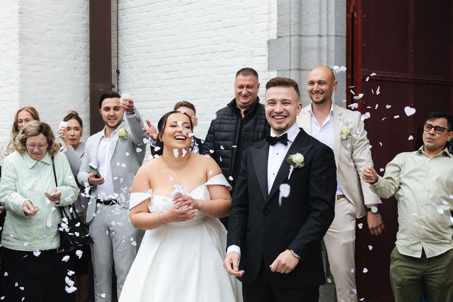 Joyful newlyweds exit church amid confetti shower, surrounded by smiling guests in elegant wedding attire.