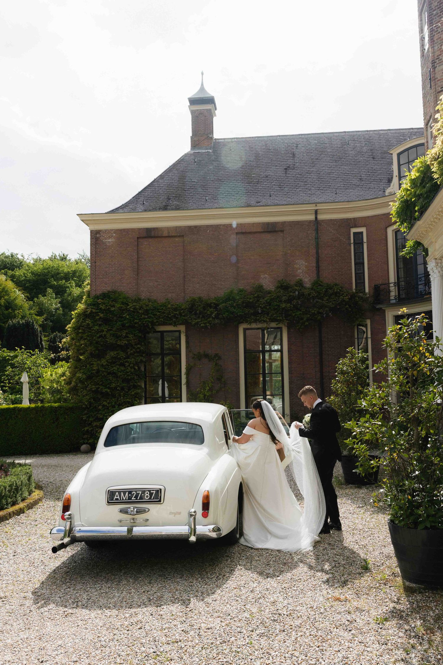 Bride and groom in elegant attire entering a vintage white classic car outside a brick manor house.