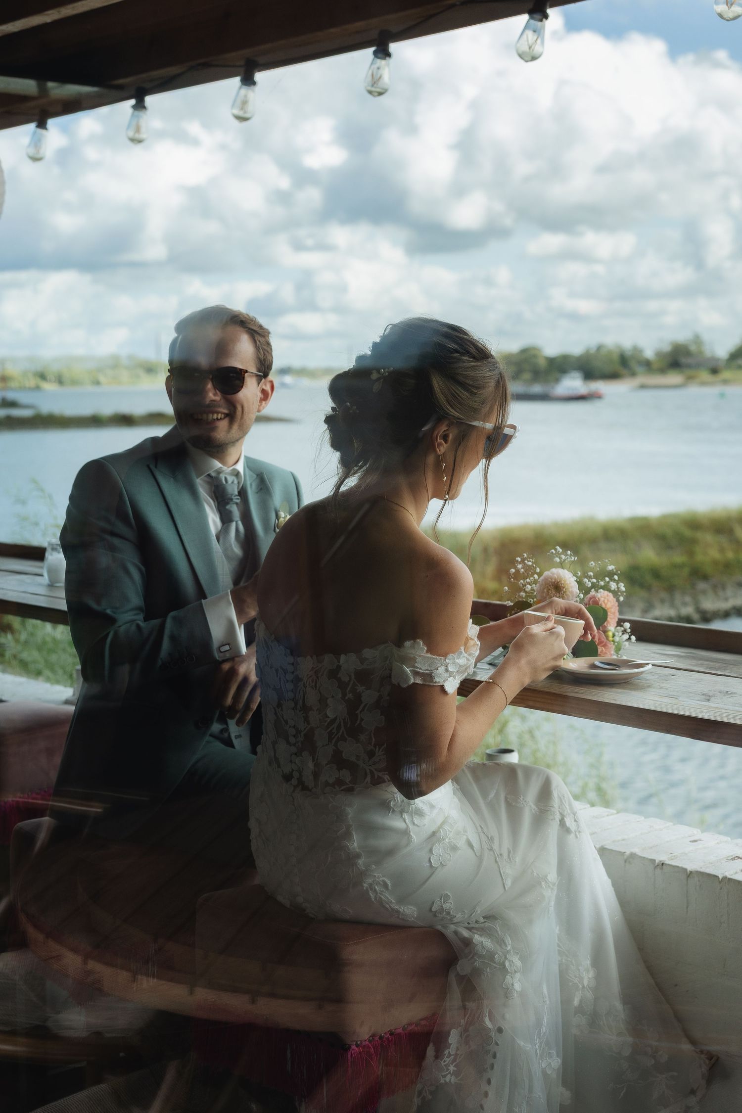 Smiling groom in gray suit and sunglasses sits with bride in lace dress at waterfront venue.