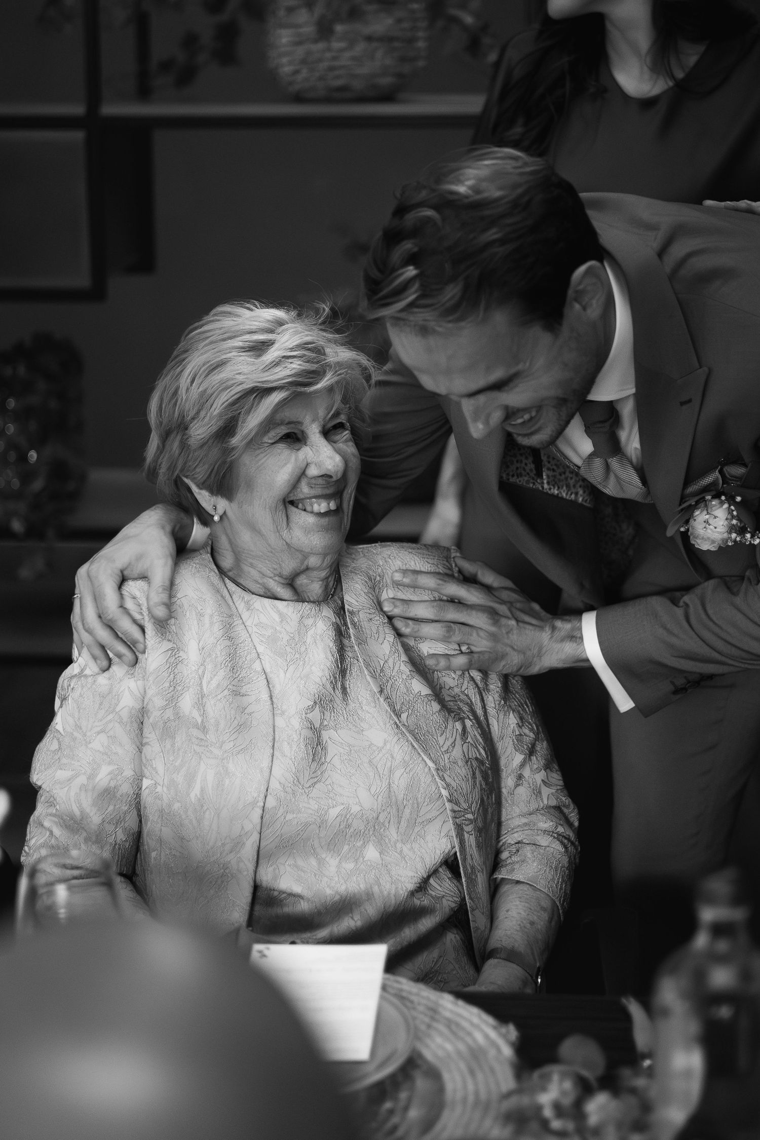 Black and white photo of an elderly woman smiling warmly as a suited groom leans in to embrace her at a wedding.