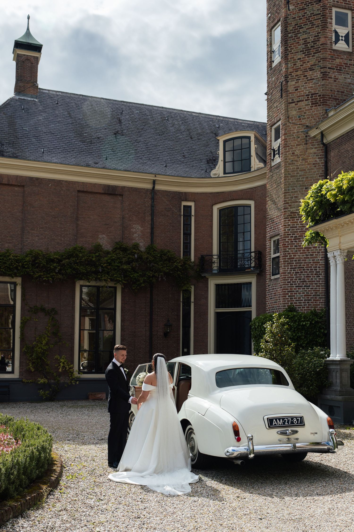 Bride and groom standing beside a classic white vintage car outside an elegant brick manor house on their wedding day.