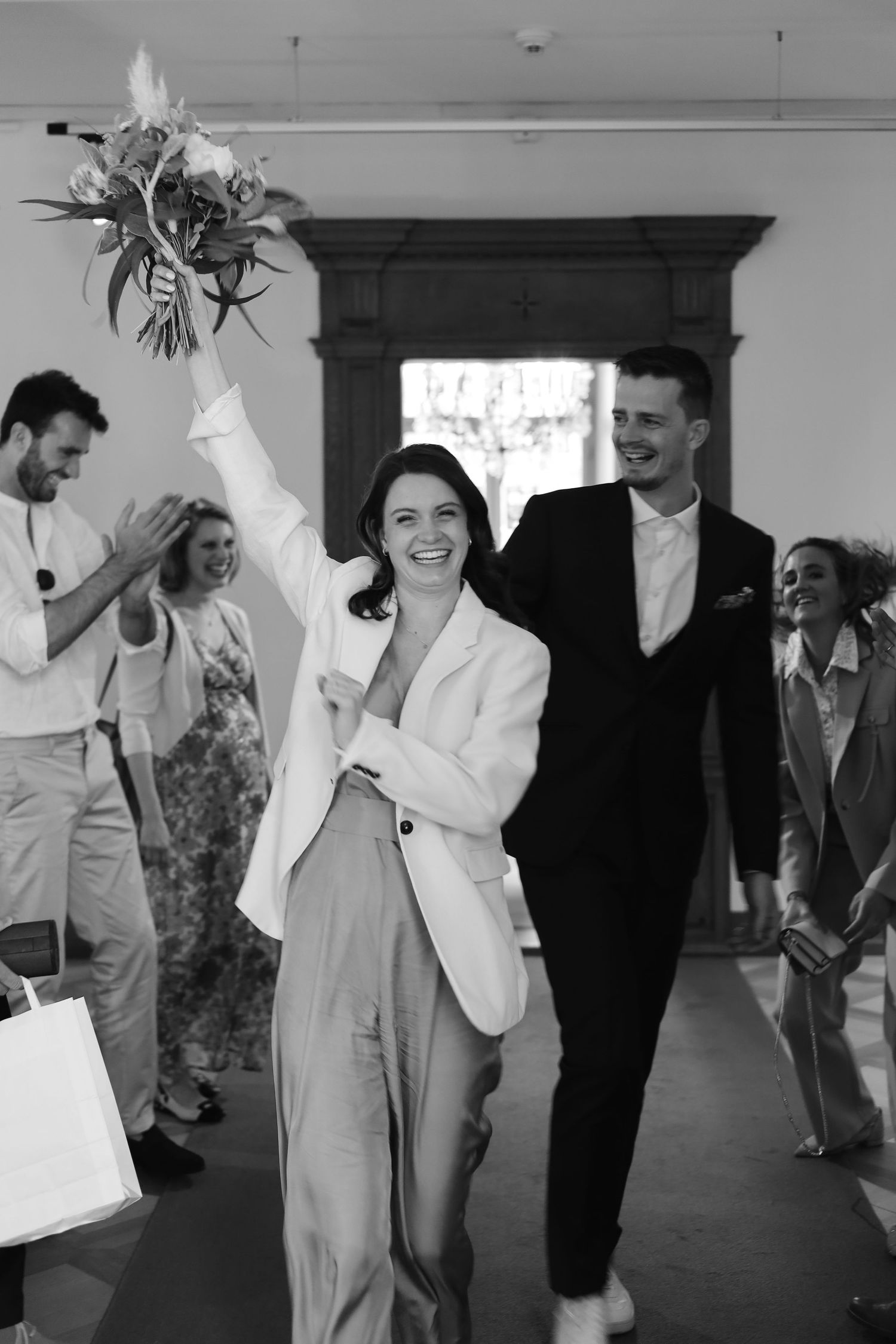 Tijdloze zwart-wit foto van een bruid en bruidegom die juichend naar buiten komen na hun ceremonie. Vrolijk en documentair beeld met gasten