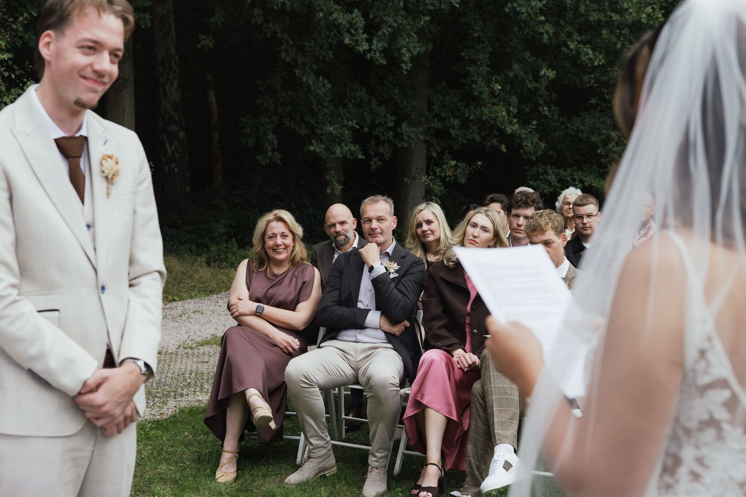 Groom in beige suit smiles as bride reads vows at outdoor wedding ceremony with seated guests.