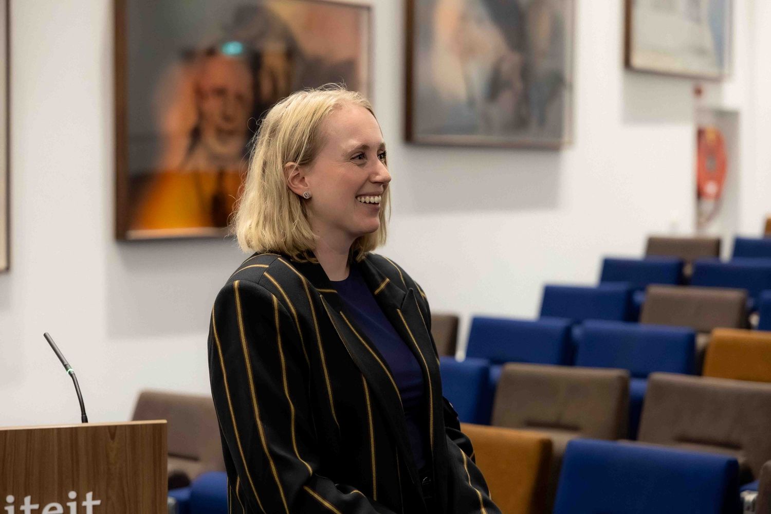 Smiling blonde woman in striped blazer standing in a formal lecture hall with portrait paintings on the wall.