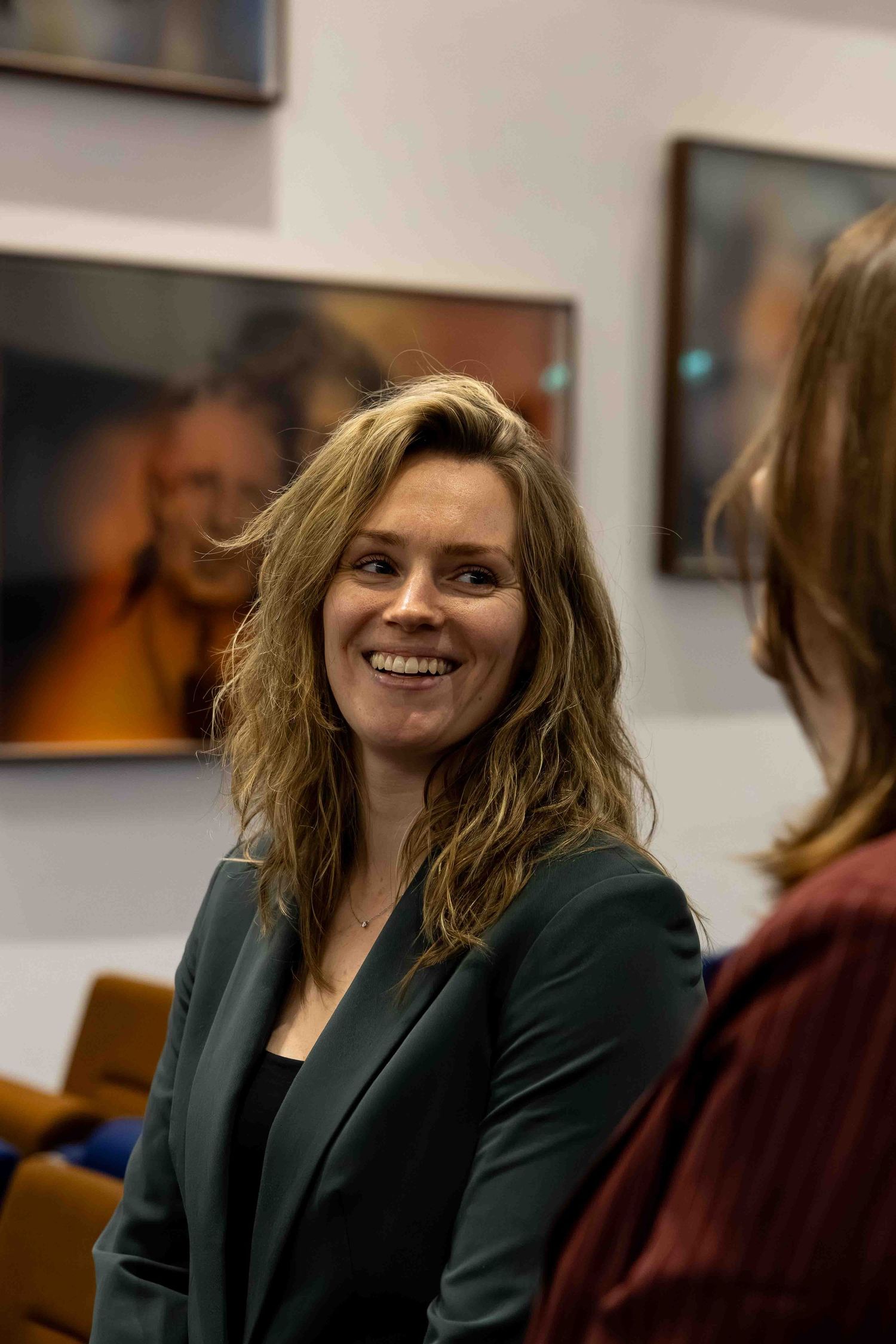 Smiling woman with wavy blonde hair in a dark blazer at an art gallery event.