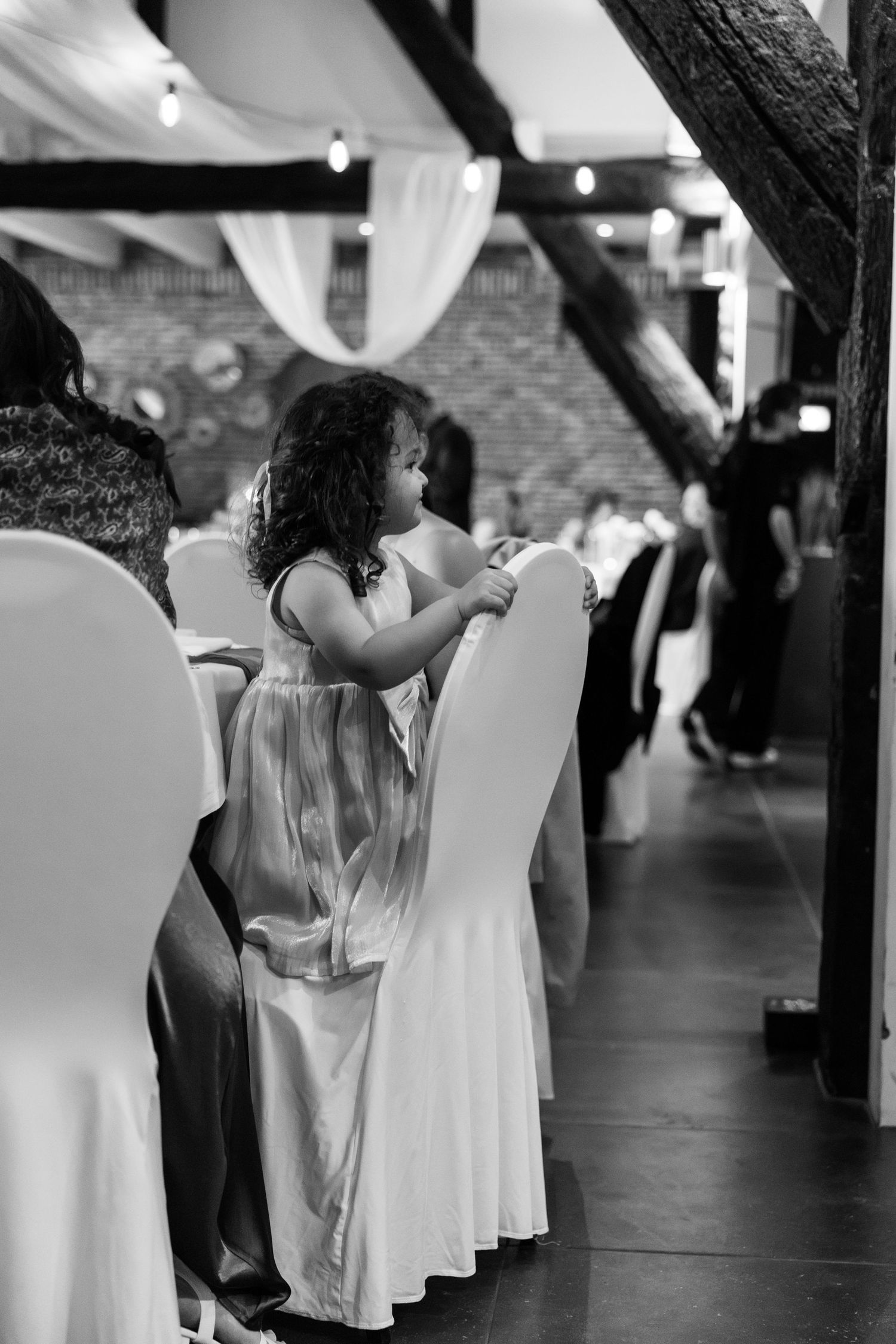 Black and white photo of a young girl with curly hair standing by chairs at an elegant indoor event.