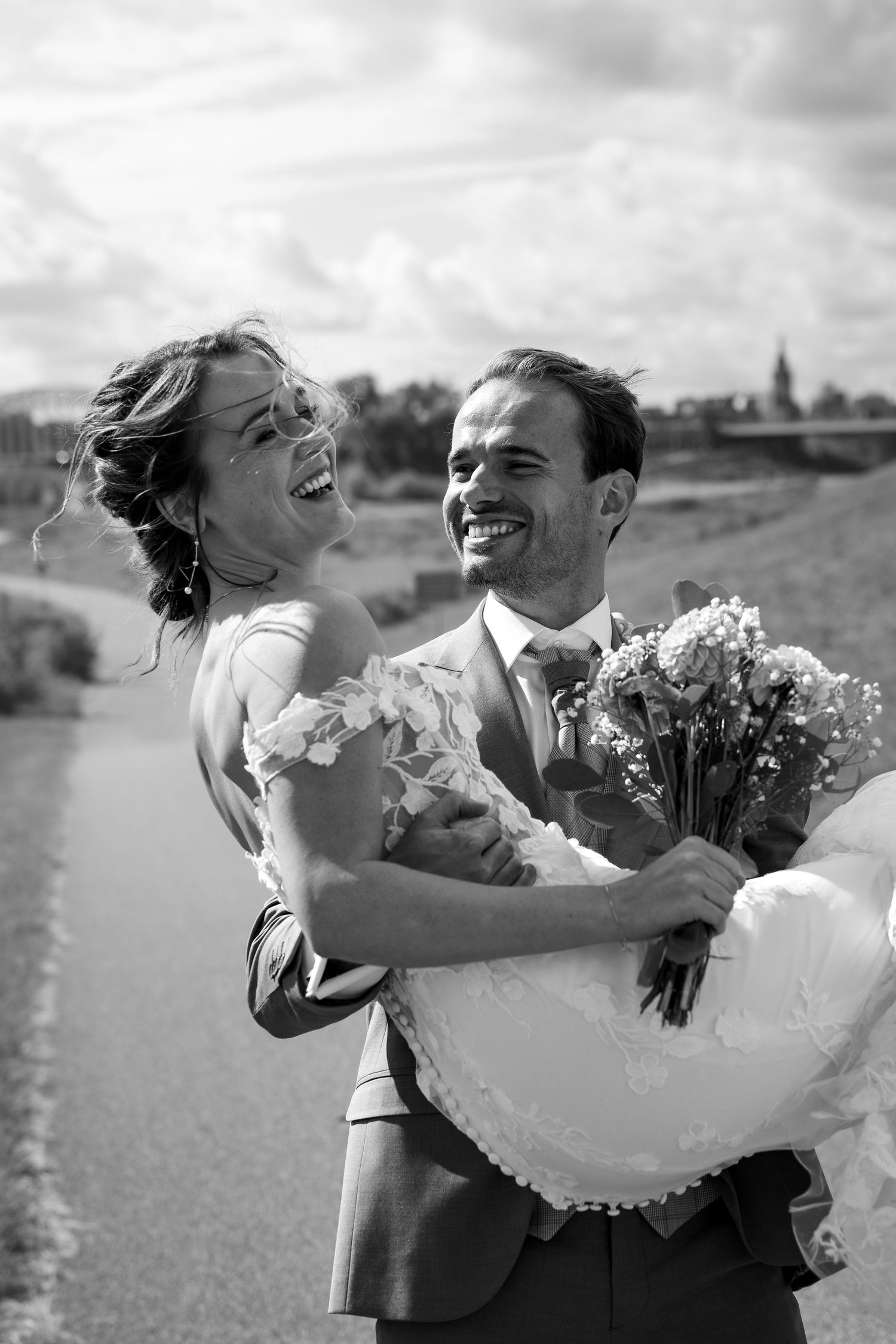 Black and white photo of a joyful bride and groom laughing together outdoors, groom holding bride and bouquet.