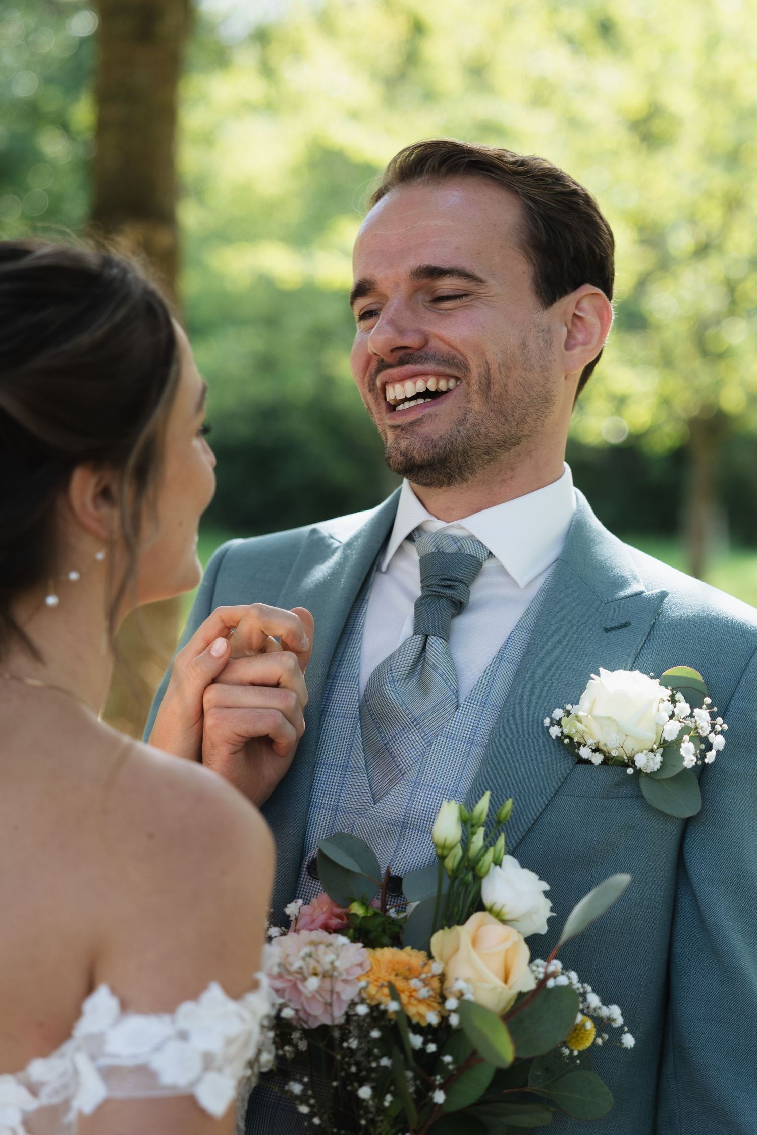 Smiling groom in light blue suit holds bride's hand outdoors, wearing boutonniere with white roses and greenery on wedding day.