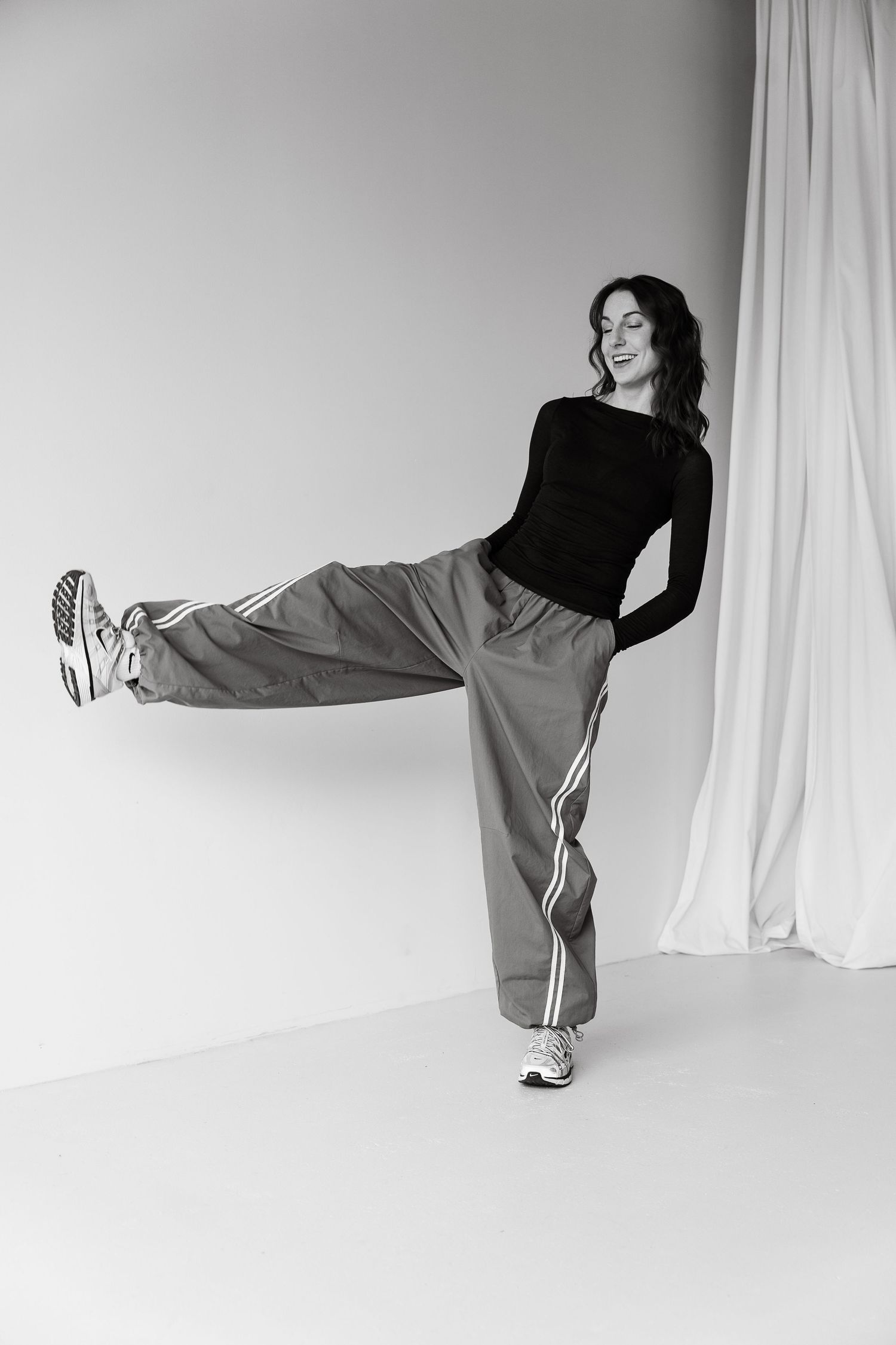 Woman in black top and wide-leg track pants kicks leg up high, posing playfully in a studio setting.