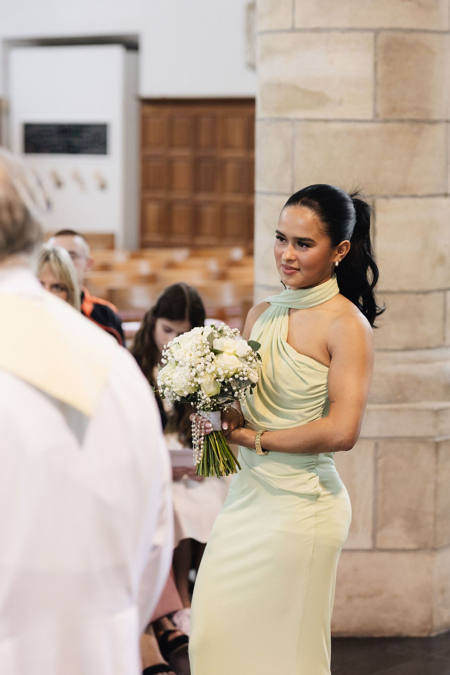 Bridesmaid in mint green halter dress holds white floral bouquet at church wedding ceremony.