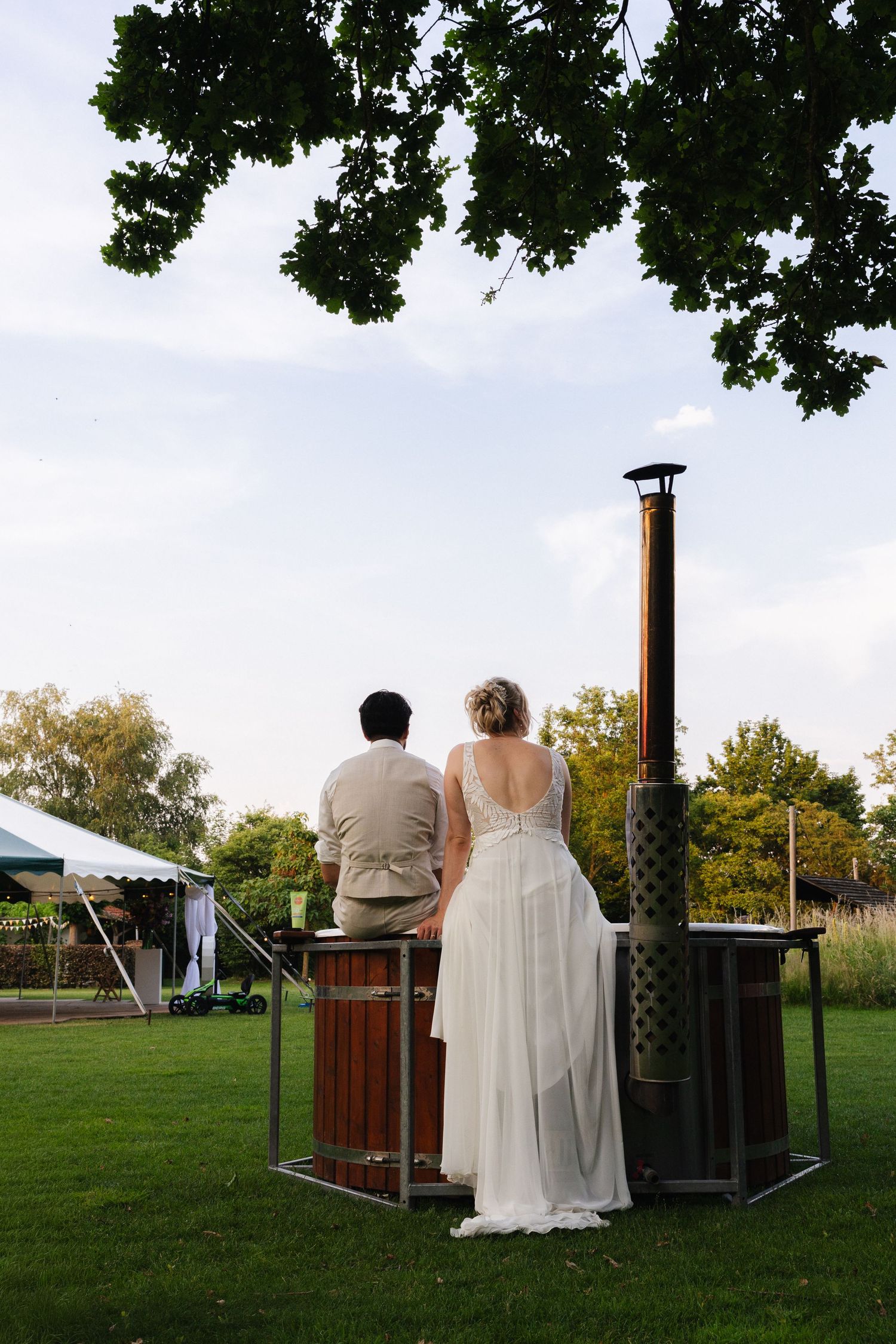 Bride in white gown and groom in beige suit stand together outdoors near a tall heater on a green lawn.