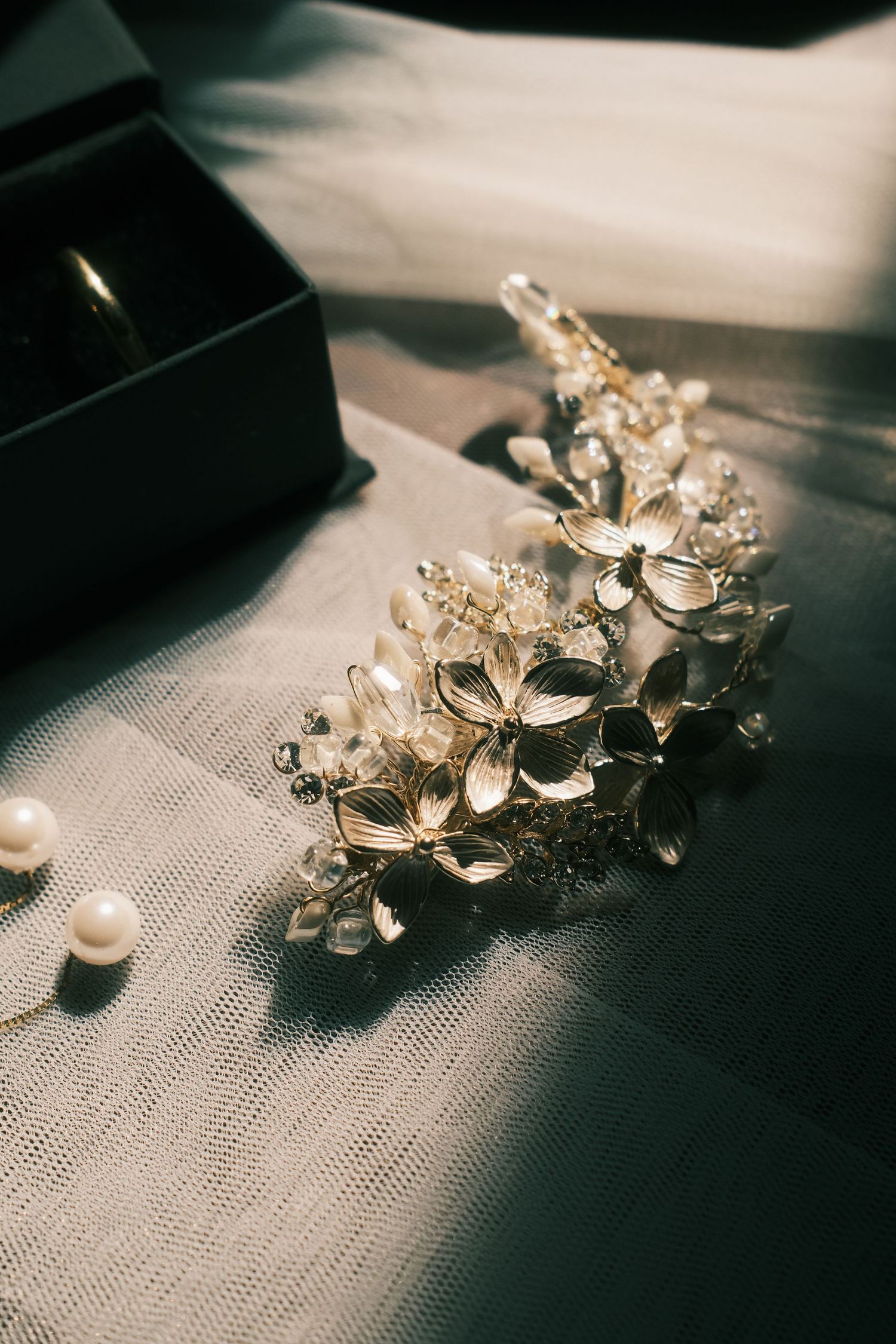 Elegant gold floral bridal hair vine with crystals beside pearl earrings and a ring box on white tulle fabric.